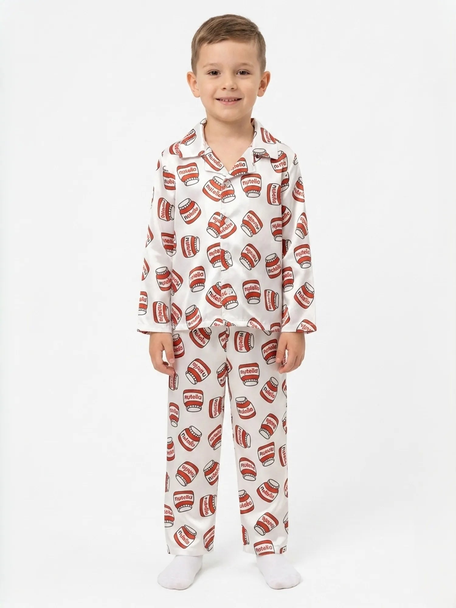 A young boy standing against a white background wearing a white satin long-sleeved night suit with a red hazelnut spread jar print.
