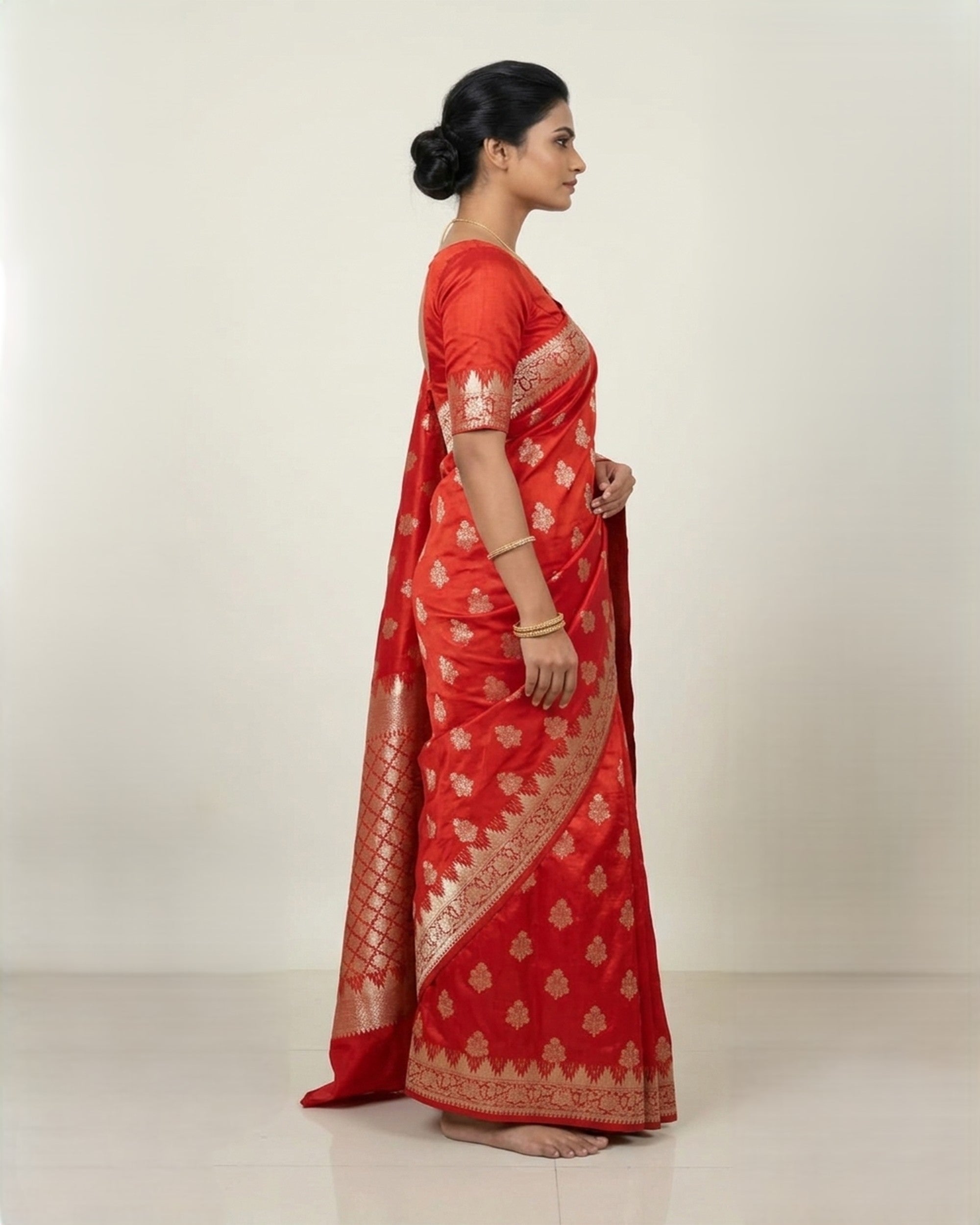 A woman wearing a vibrant red Banarasi silk saree with intricate gold zari floral patterns and a broad, decorative border, posed against a plain white background.