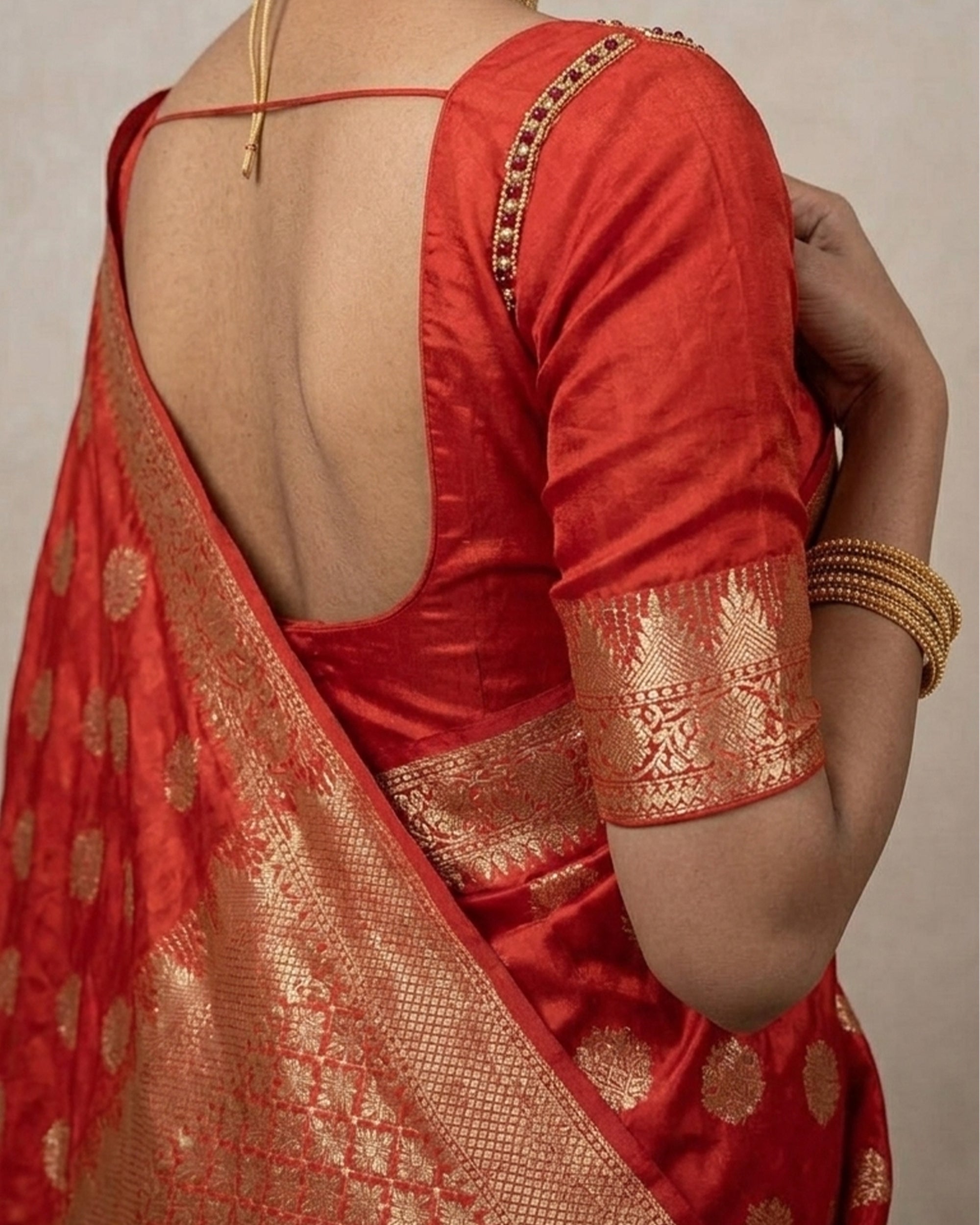 A woman wearing a vibrant red Banarasi silk saree with intricate gold zari floral patterns and a broad, decorative border, posed against a plain white background.