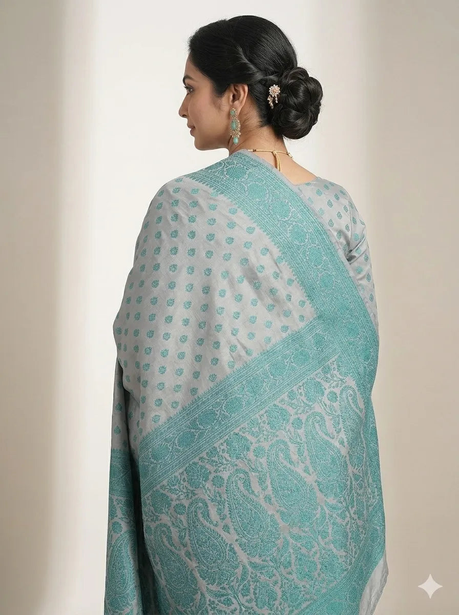 Back view of a woman wearing a grey & teal blue woven Banarasi silk saree, showcasing the heavy teal Zari pallu with intricate paisley and floral artistry, paired with an elegant braided bun and traditional earrings.