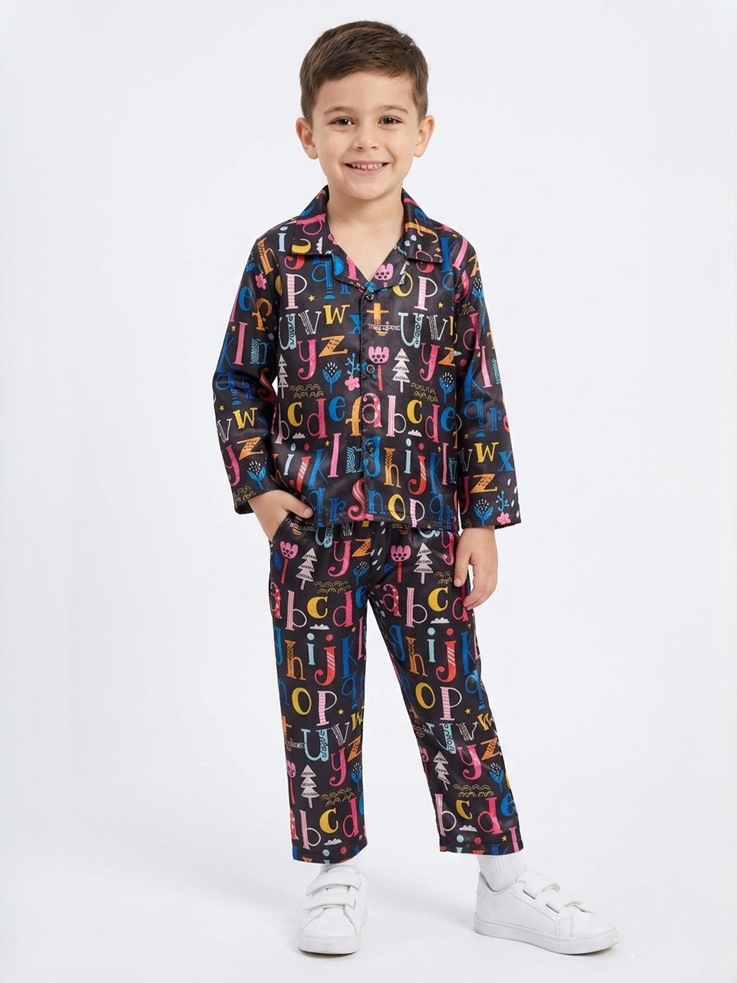 A young boy standing against a white background wearing a Black satin night suit with a multicolored alphabet print.