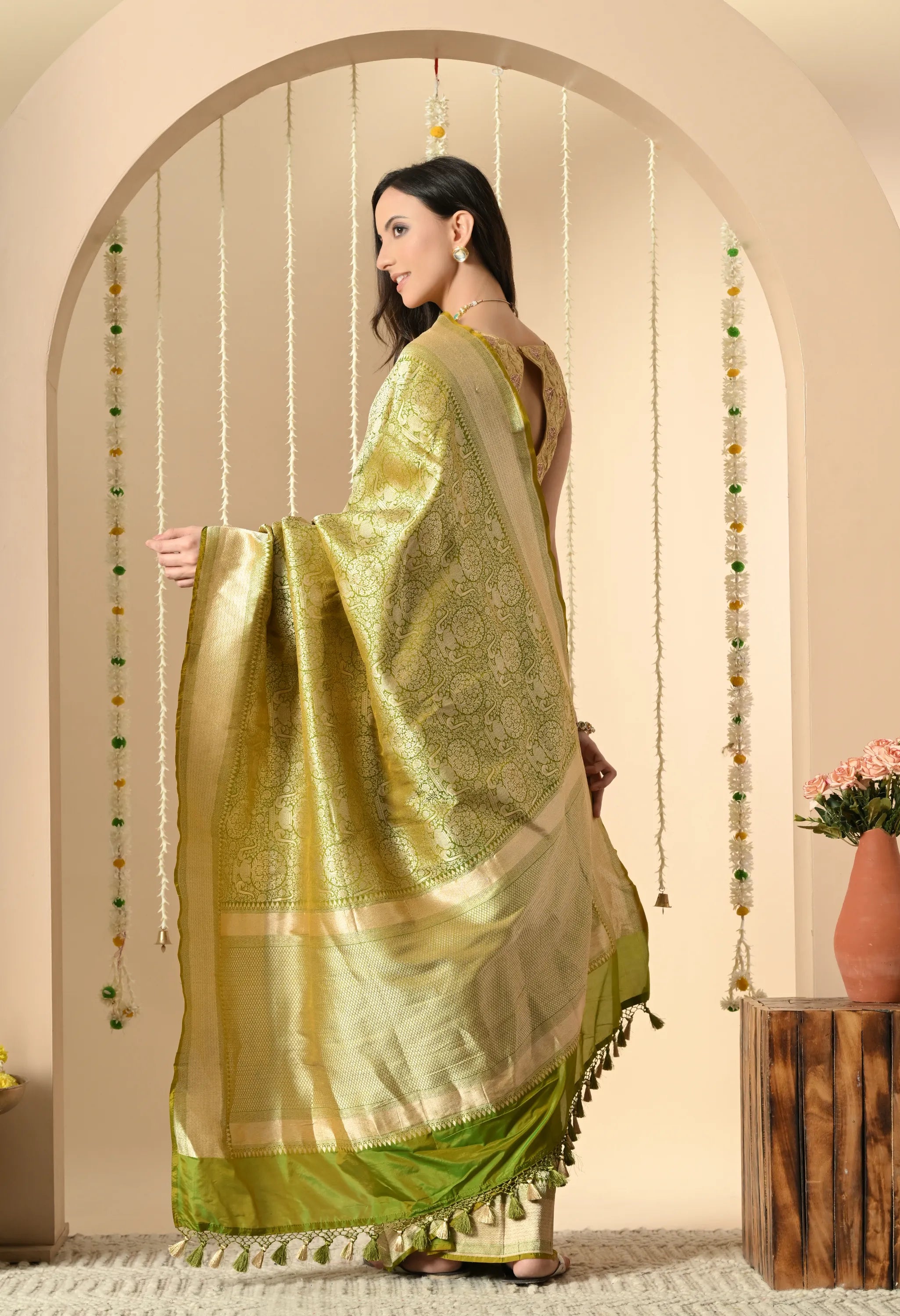 A model is standing and posing in a Silk Mark Certified traditional Indian Katan silk saree from The Weavers Shop by Raavi. The saree is a bright, shimmering lime green with intricate gold zari work (brocade) across the body and pallu (decorative end). The borders are also adorned with gold zari work. She is wearing a sleeveless gold blouse and traditional jewelry, including a necklace and earrings. She is standing in an archway decorated with strings of marigold and jasmine flowers.