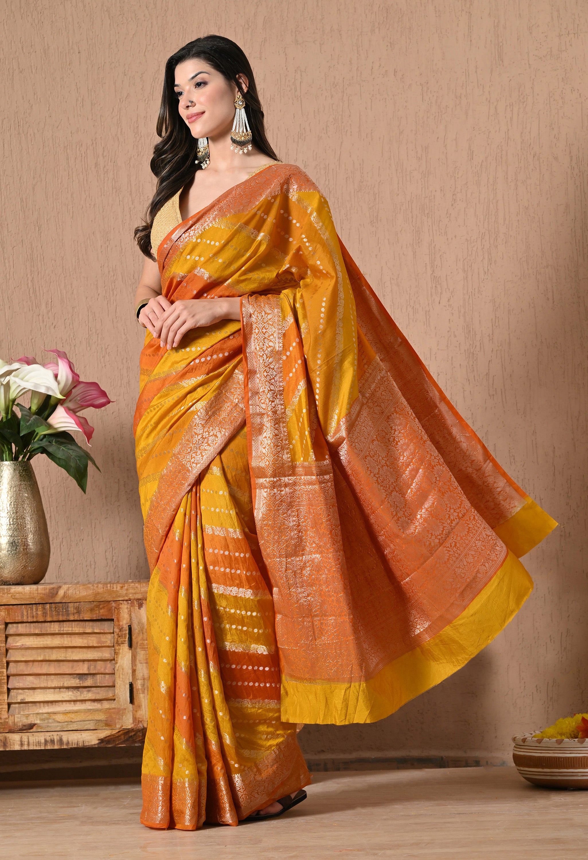 A model stands in a three-quarter pose wearing a Rangkat Banarasi silk saree featuring a warm orange and yellow sunset-toned palette. The saree is embellished with elaborate golden zari work on the borders and pallu, featuring traditional geometric and fine woven motifs. The body of the saree displays diagonal Rangkat stripes decorated with delicate silver-toned polka dots. The look is styled with a gold sequined blouse, statement pearl-drop earrings, and a metallic gold cuff.