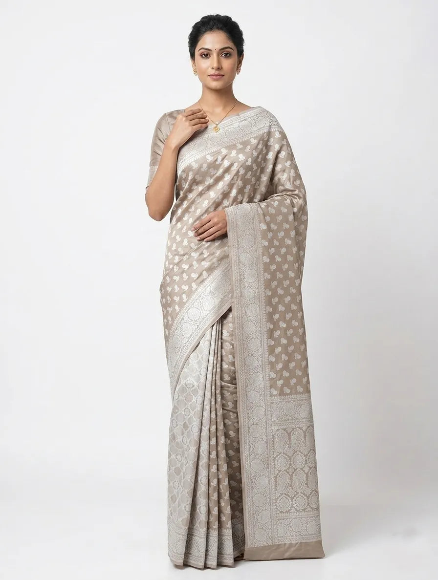Model draped in a beige and silver silk blend Banarasi saree featuring an intricate woven zari floral design and a broad ornamental border.