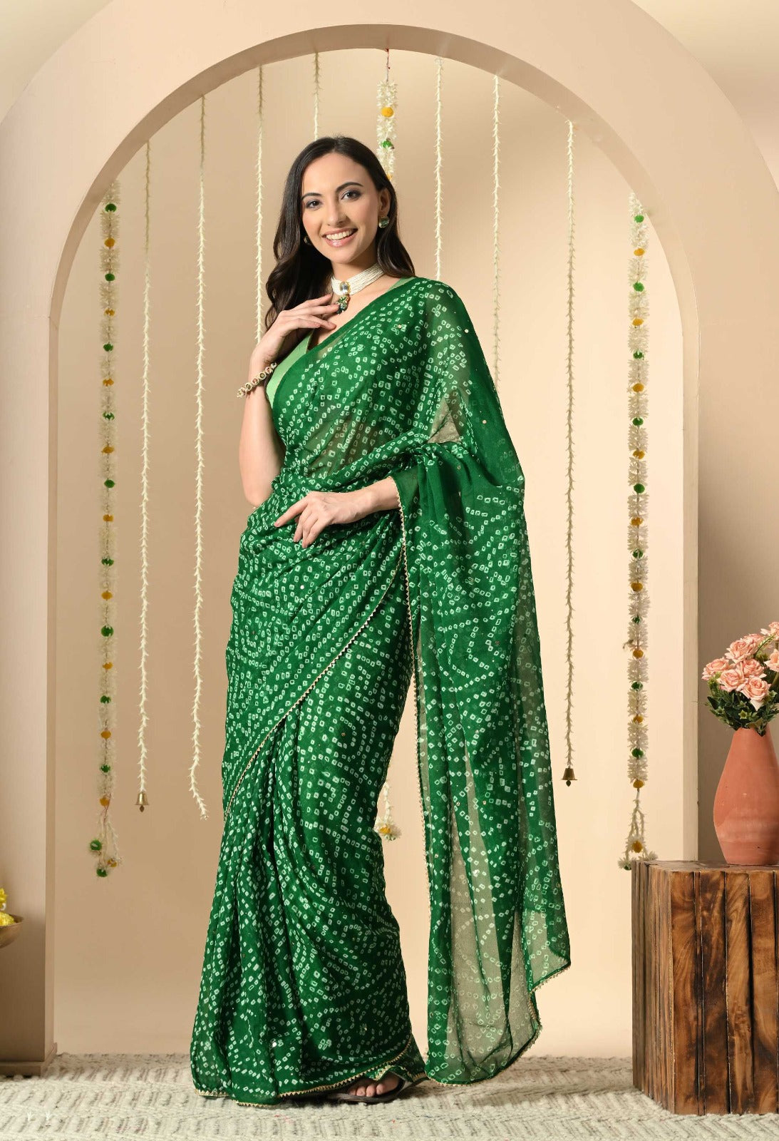 The image displays a woman elegantly wearing a vibrant green Bandhani saree. This traditional Indian garment features a distinctive tie-dye pattern spread uniformly across the fabric. The saree is crafted from a lightweight, fluid material such as georgette or chiffon, allowing it to drape gracefully around the body. It is paired with a matching green sleeveless blouse, creating a unified and monochromatic look.