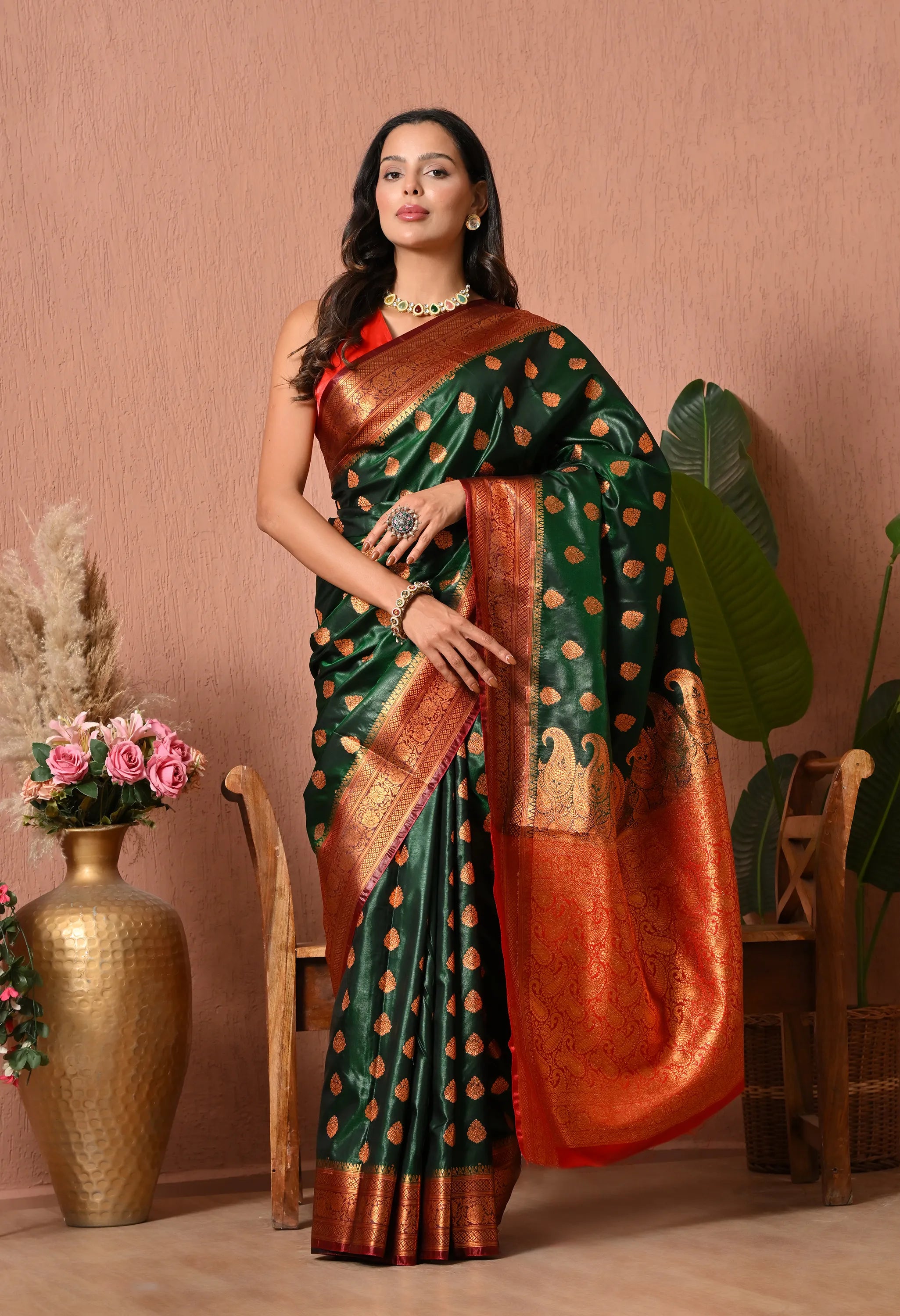 A woman posing in a rich bottle green silk blend saree featuring allover gold zari motifs and a dramatic crimson red contrast border and pallu with intricate traditional patterns.