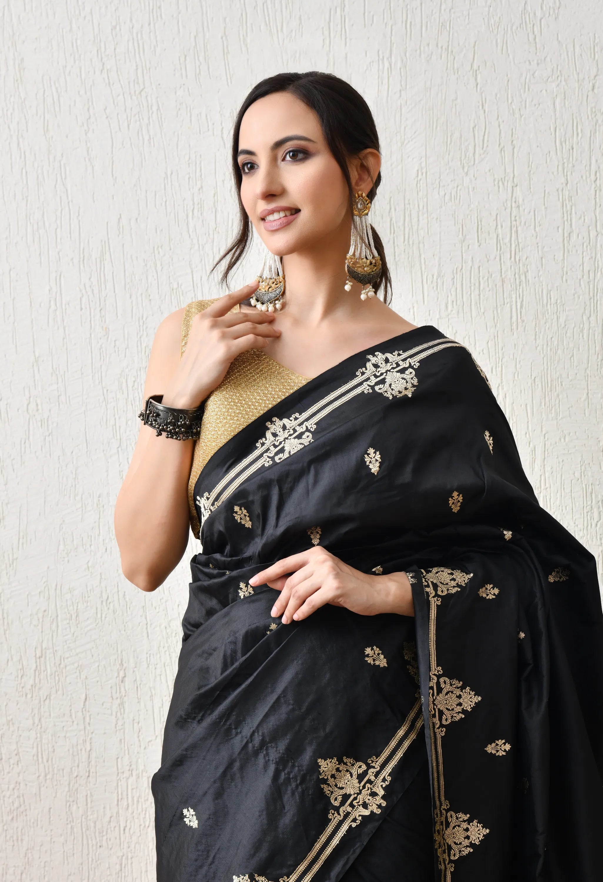 A smiling woman in a rear-facing pose is wearing a black Silk Mark Certified Banarasi saree from The Weavers Shop by Raavi . The saree's rich gold zari Karuwa weave is highly visible, featuring large circular floral motifs across the pallu, a thick gold border, and scattered gold bootis on the body. She is wearing a gold blouse with a delicate back tie, large gold statement earrings, and dark bangles. She stands indoors next to a rustic wooden cabinet against a textured white wall