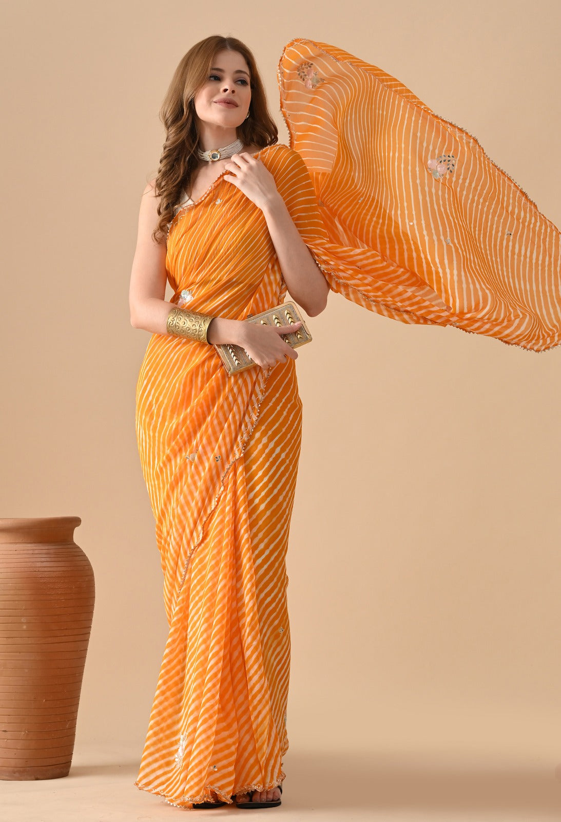 A woman poses in a vibrant yellow-orange Leheriya saree with thin, diagonal white wave stripes. She pairs it with a modern white one-shoulder blouse, a wide metallic gold cuff bracelet, a layered pearl choker necklace, and a small gold textured clutch bag. The fabric of the saree is lightweight and flows gracefully in the air against a minimalist, solid beige background next to a rustic clay pot.