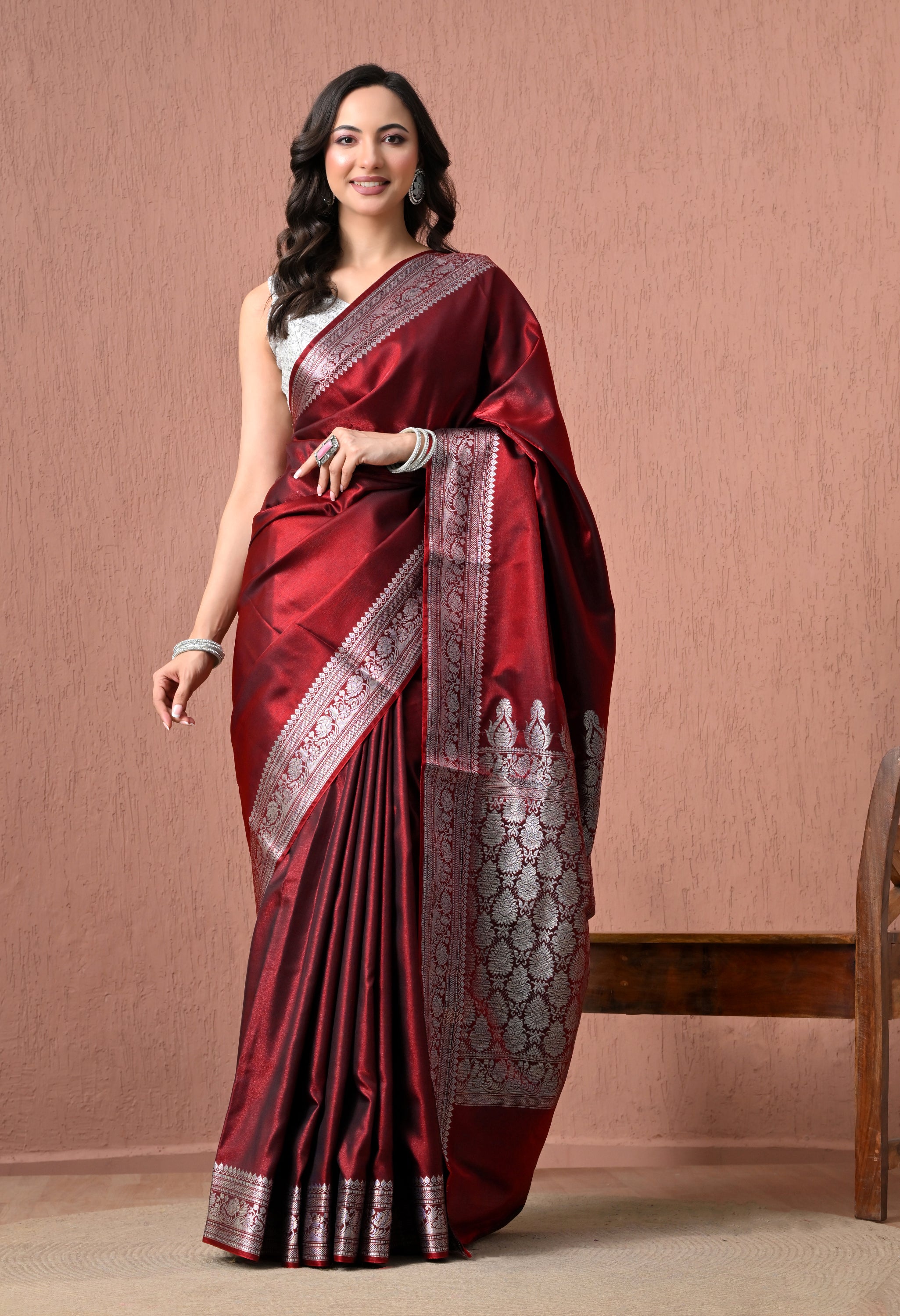 A studio portrait of a woman modeling a luxurious, deep maroon pure silk Banarasi saree. The saree features a rich, reflective texture with intricate silver zari floral work along the wide borders and pallu. The model is smiling, wearing a sleeveless white and silver textured blouse, silver statement earrings, and matching bangles, standing against a textured peach-colored wall background next to a wooden chair.
