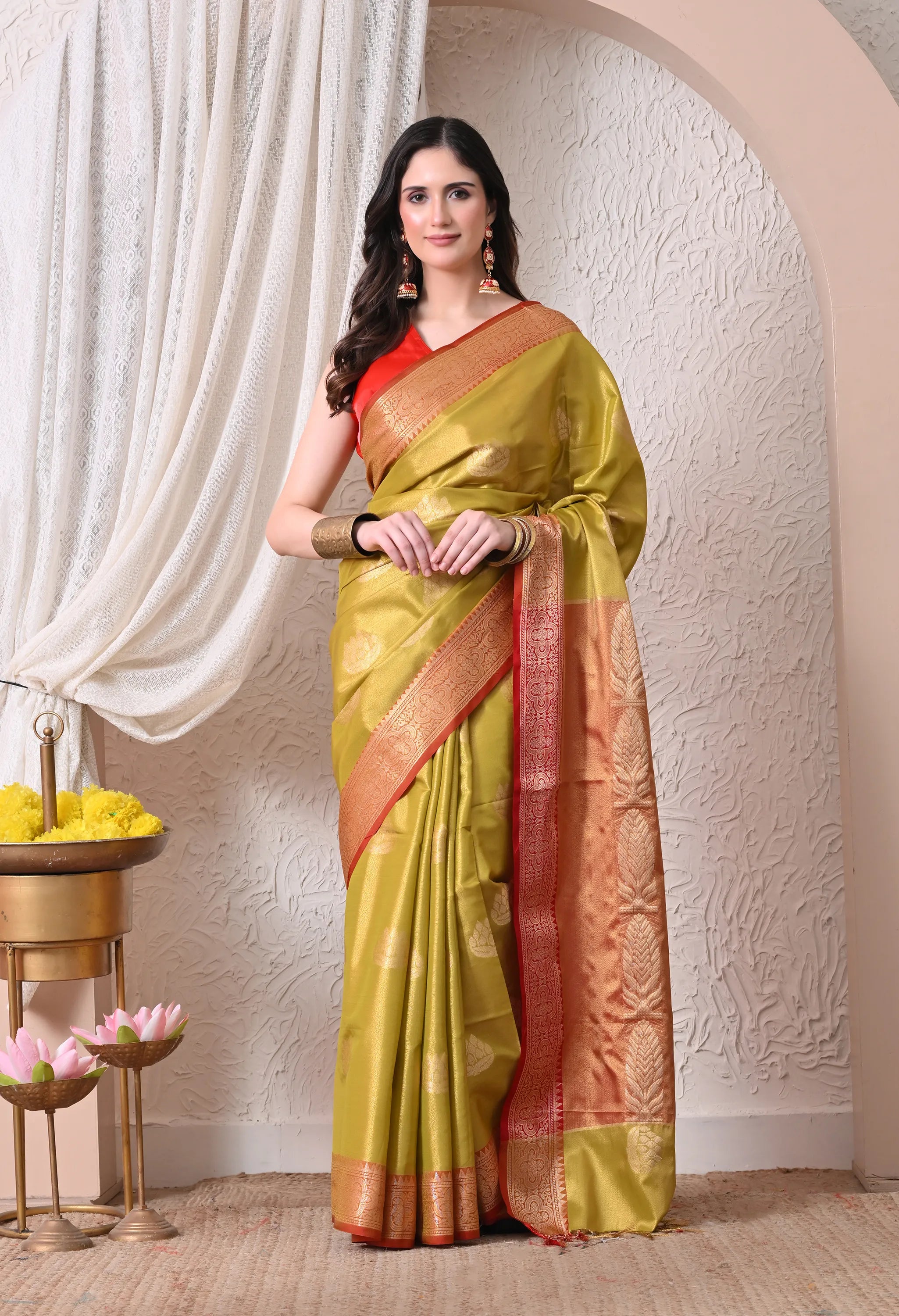 A woman stands posing in an elegant olive-green silk blend saree featuring a wide crimson-red border with intricate gold zari embroidery and a matching red blouse.