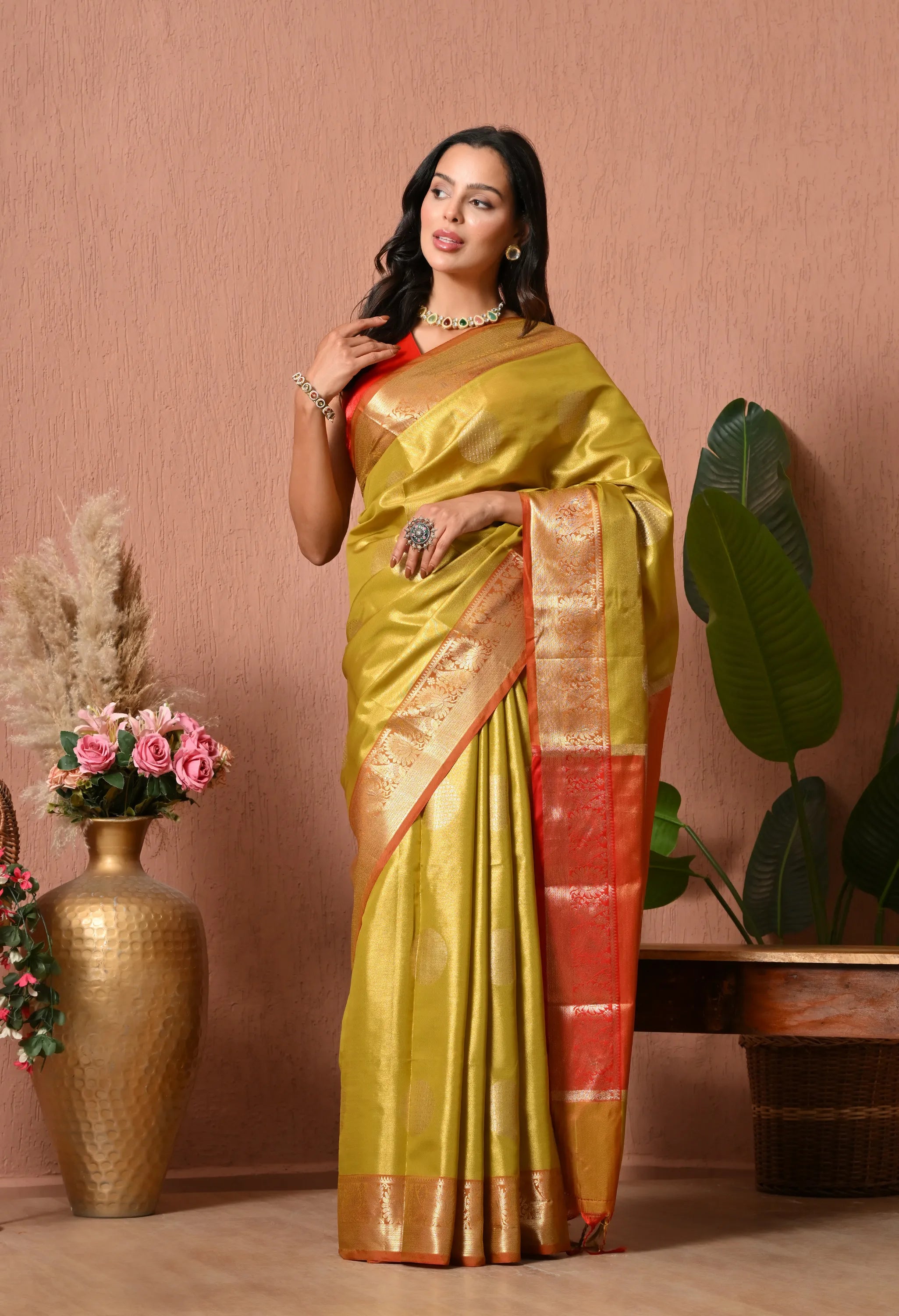 A woman wearing an elegant olive green silk blend saree featuring a wide, ornate red and gold contrast zari border. She is accessorized with traditional jewelry against a warm textured background.