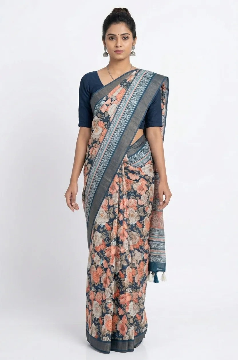 A woman standing in a deep teal silk blend saree featuring a dense floral print in peach and white, styled with a solid navy blue blouse and silver earrings.
