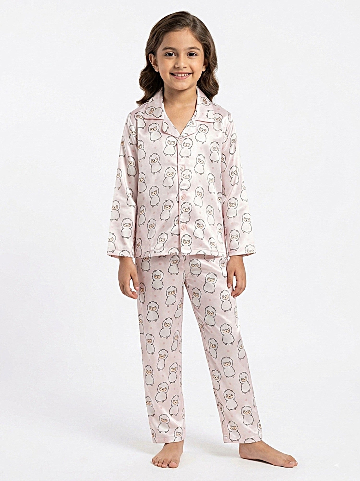 A young girl smiling while posing in a two-piece pastel pink satin night suit printed with cute cartoon animal faces, featuring a button-up long-sleeve shirt and matching pajama pants against a white background.