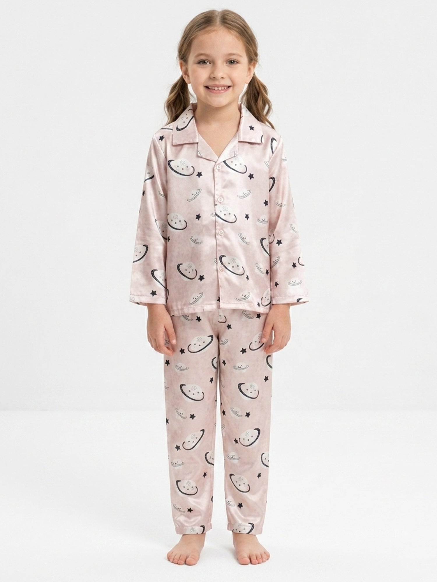 A smiling young girl with two pigtails posing in a light pink satin night suit set decorated with moon, star, and planet prints against a solid white background.