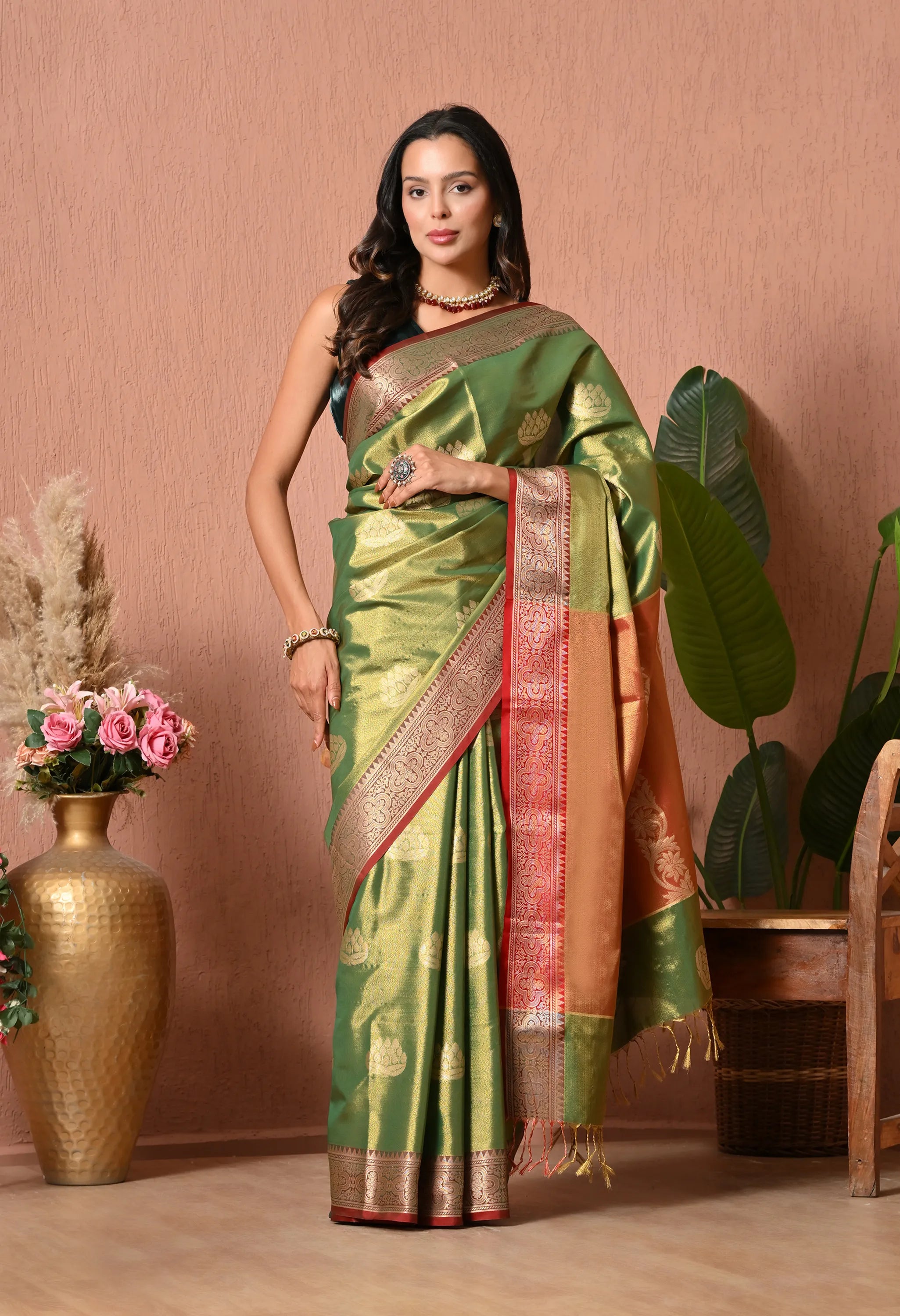 A woman posing elegantly against a dusty pink wall in an olive green silk blend saree with gold lotus motifs and a multi-toned crimson and rust zari pallu.