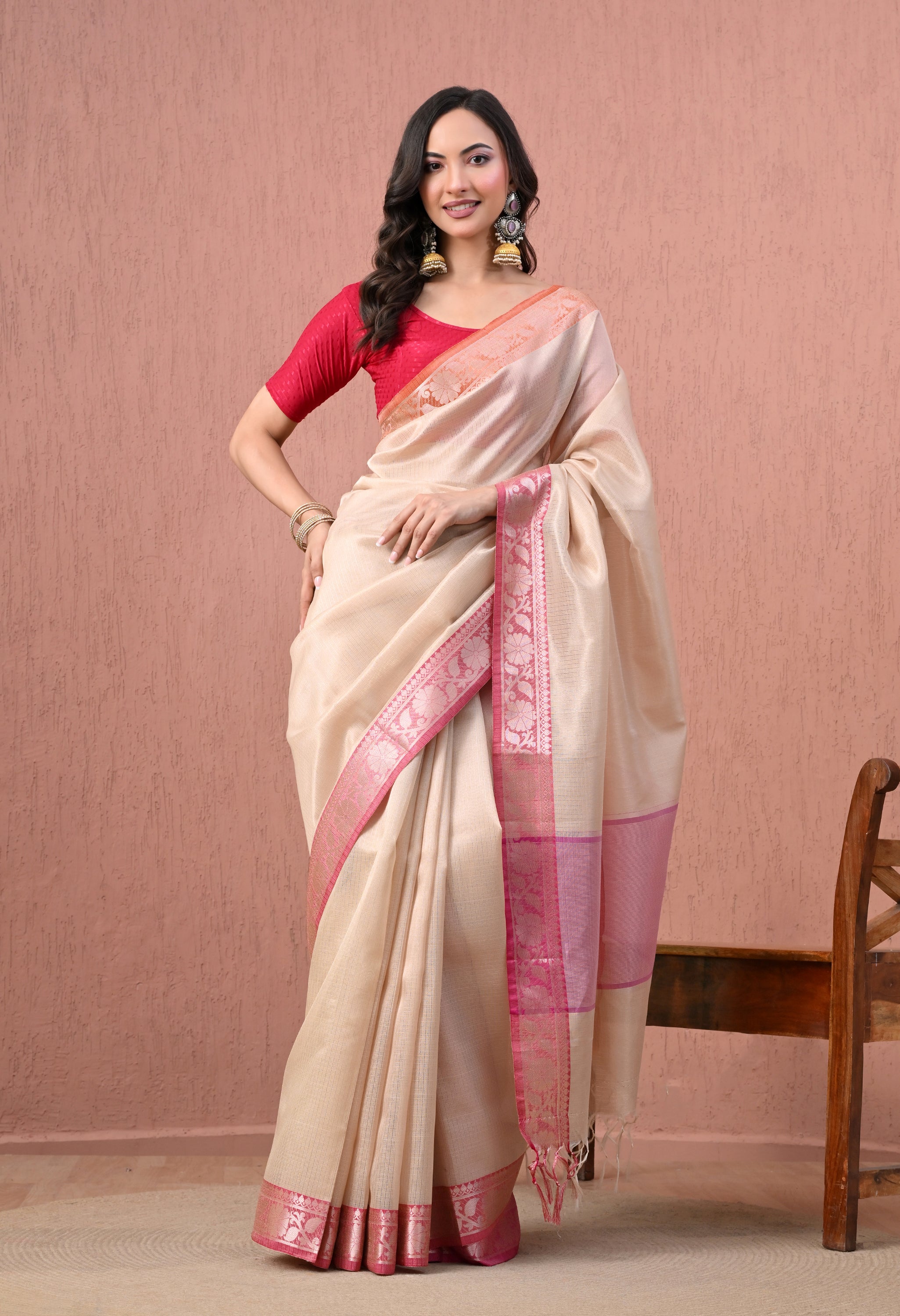 A full-length photo of a woman posing in an elegant cream-colored pure cotton handloom saree. The saree features a bright Rani pink and zari floral border with a matching pink pallu and a solid pink elbow-length blouse.
