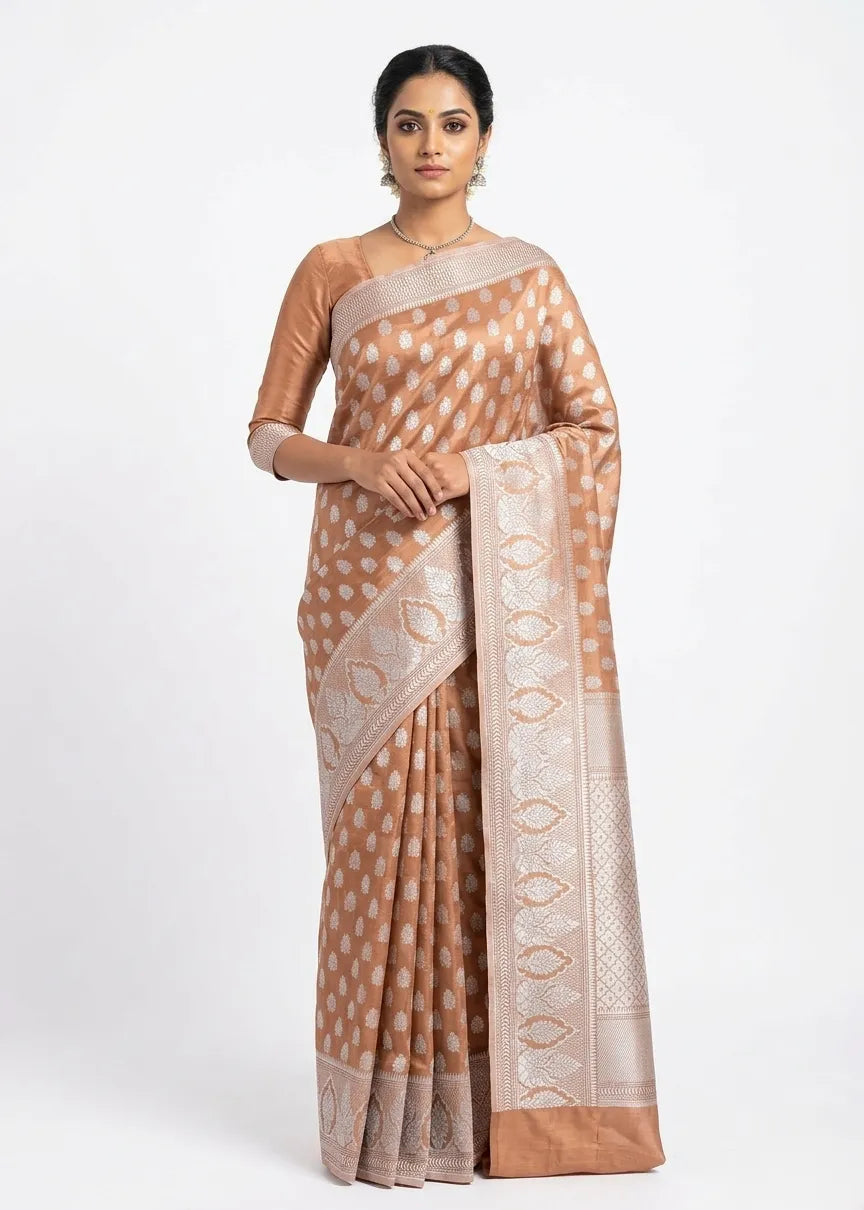 A woman posing in a peach and silver silk blend Banarasi saree with intricate zari floral motifs and a broad decorative border against a white background.

