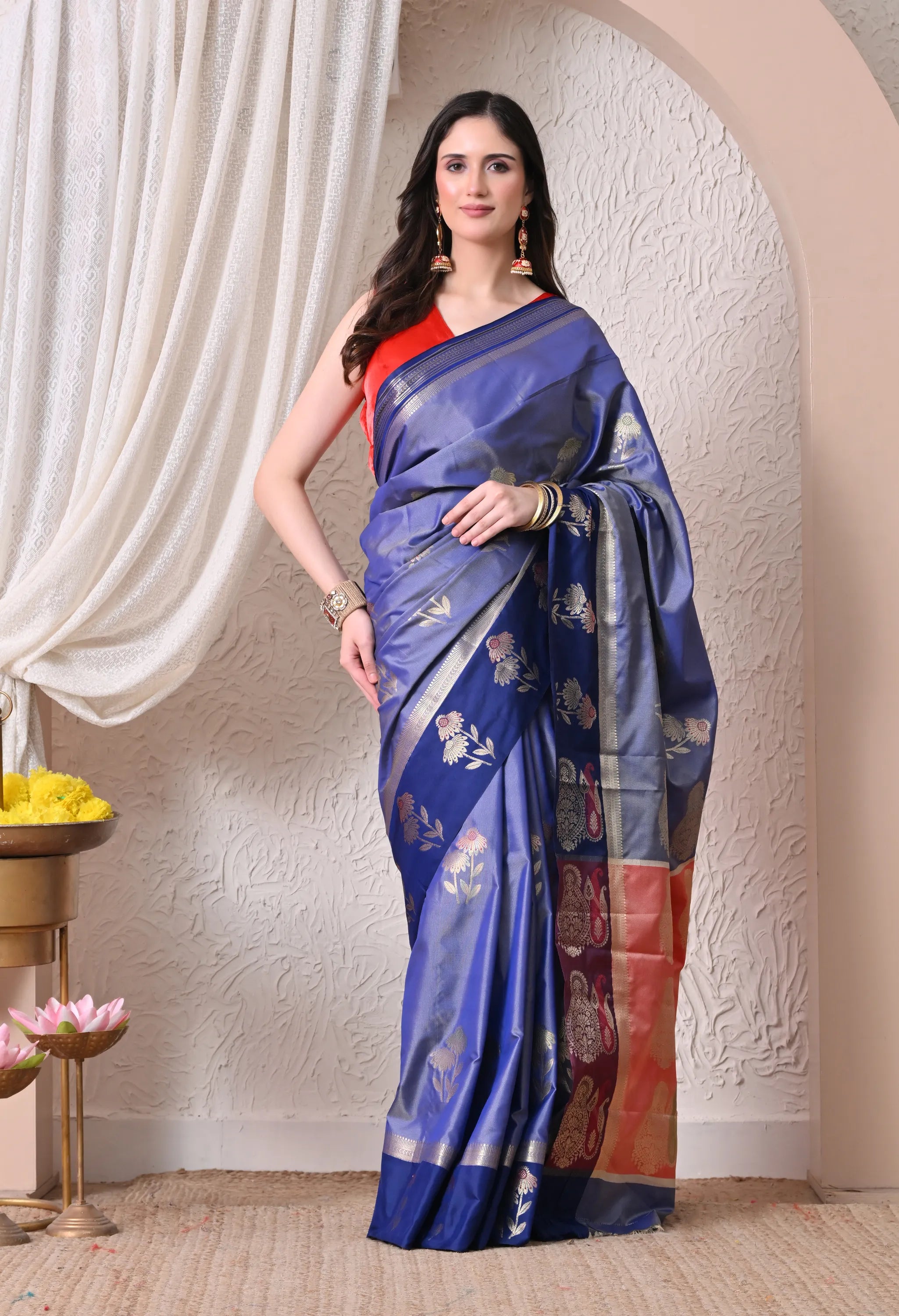 A woman poses in a vibrant royal blue silk blend saree featuring a contrasting navy blue pallu with silver floral zari motifs and a bright coral-red blouse.
