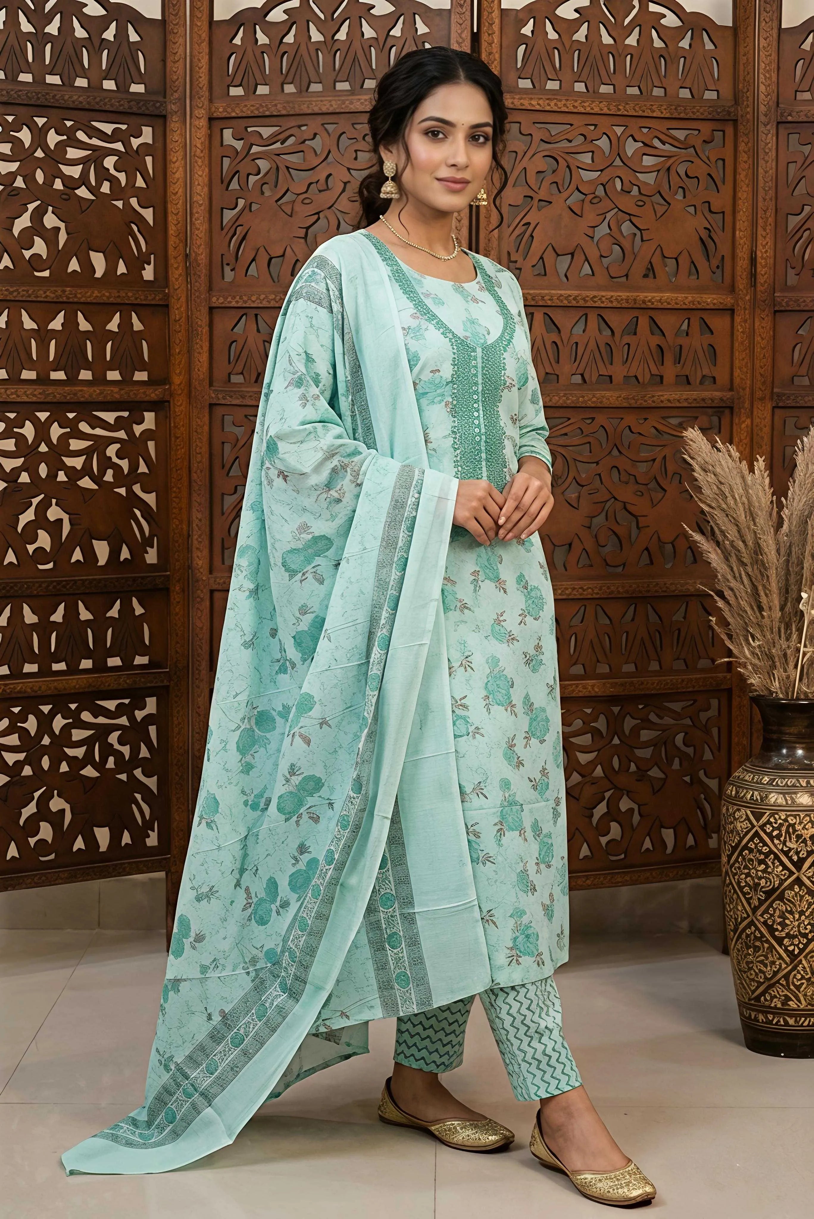 
A woman wearing a sea green and sage floral printed unstitched cotton suit. The outfit features an embroidered round neckline, a matching printed dupatta with a patterned border, and floral printed trousers, styled in front of a wooden carved screen.