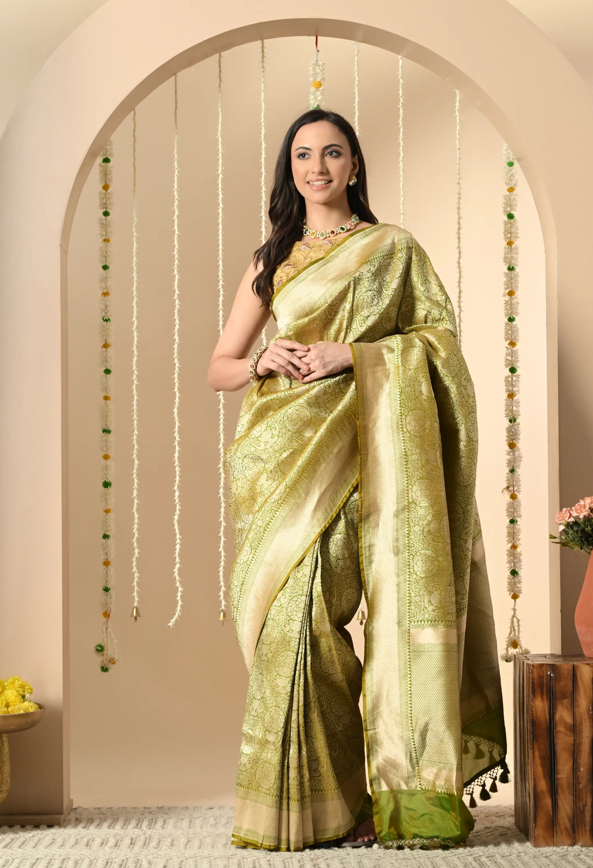 A model is standing and posing in a Silk Mark Certified traditional Indian Katan silk saree from The Weavers Shop by Raavi. The saree is a bright, shimmering lime green with intricate gold zari work (brocade) across the body and pallu (decorative end). The borders are also adorned with gold zari work. She is wearing a sleeveless gold blouse and traditional jewelry, including a necklace and earrings. She is standing in an archway decorated with strings of marigold and jasmine flowers.