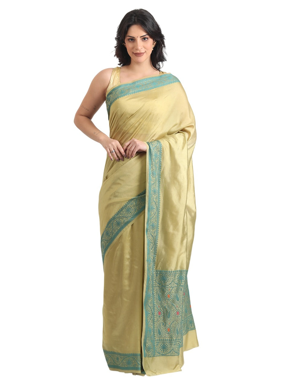 A woman is standing against a plain white background, showcasing a light olive-gold silk saree with a smooth, satin finish. The saree features a contrasting turquoise-blue border and an intricately detailed pallu, both woven with traditional paisley and floral patterns. She wears a matching sleeveless gold blouse and has her dark hair styled down, posing with one hand gently resting on the draped fabric.