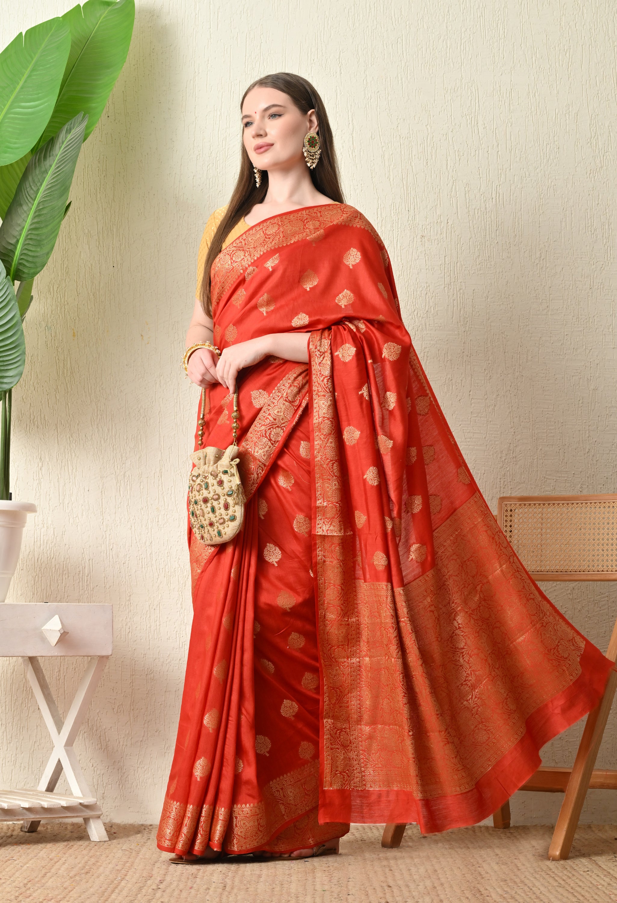 A side-profile view of a woman modeling a vibrant red Banarasi silk saree from The Weavers Shop by Raavi. The image captures the elegant drape of the fabric, highlighting the wide, ornate gold zari border and the repeating Tilfi embroidery that extends across the length of the saree. The red silk is paired with a gold-textured blouse, showcasing a sophisticated look suitable for wedding events.