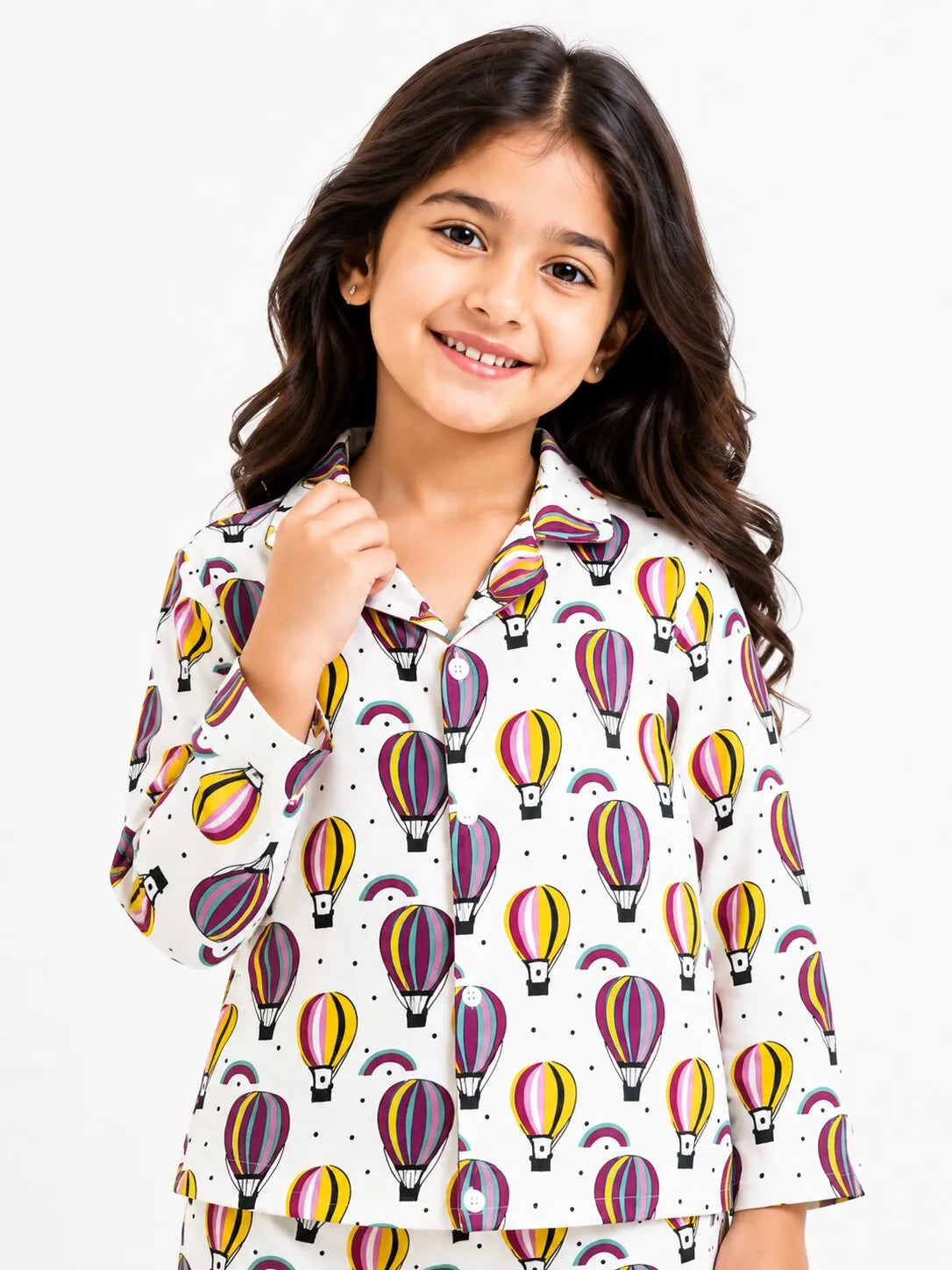 A young girl with long, wavy dark hair smiling brightly while holding the collar of her pajama top. She is wearing a white long-sleeve pajama shirt featuring a vibrant, colorful repeating pattern of hot air balloons in shades of yellow, purple, pink, and orange, set against a plain white background.