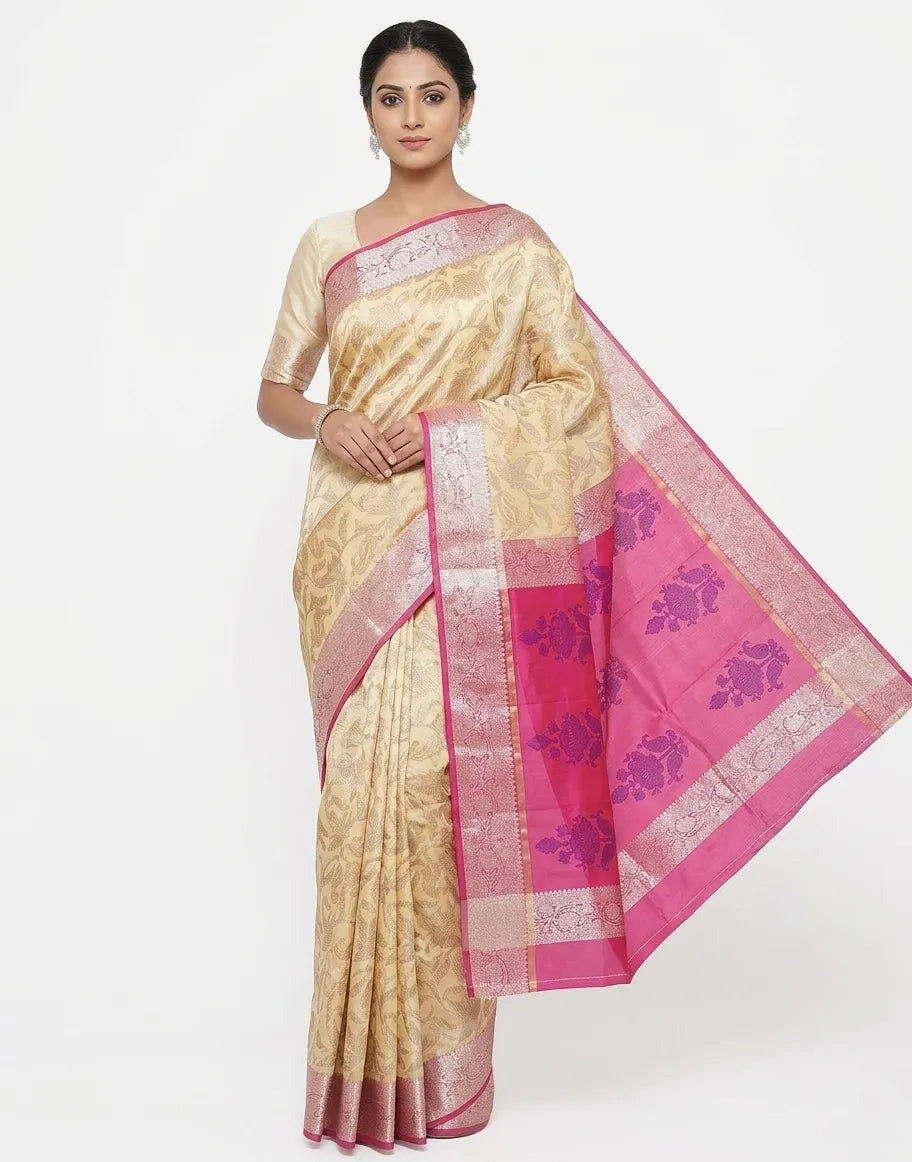 An Indian woman in a cream silk blend saree with tonal floral weaving and a bold pink zari border. She wears a matching cream blouse and silver jewelry against a plain white background.
