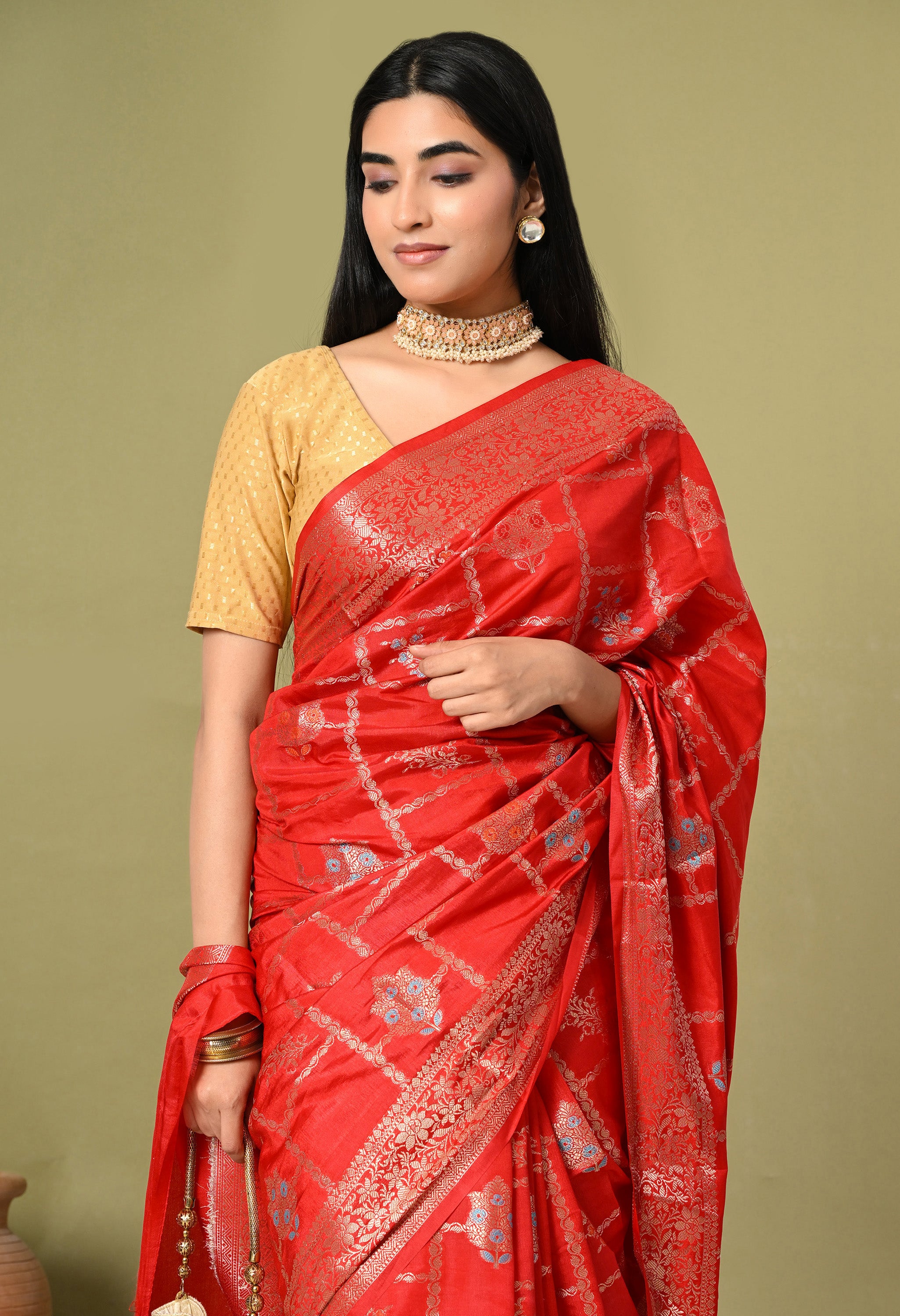 A medium close-up shot focusing on the rich texture and detailed Tilfi work of a saree from The Weavers Shop by Raavi. The red silk fabric features a shimmering gold zari grid and finely woven floral motifs. The shot highlights the craftsmanship of the gold border and the model's coordinated jewelry, showcasing the premium quality