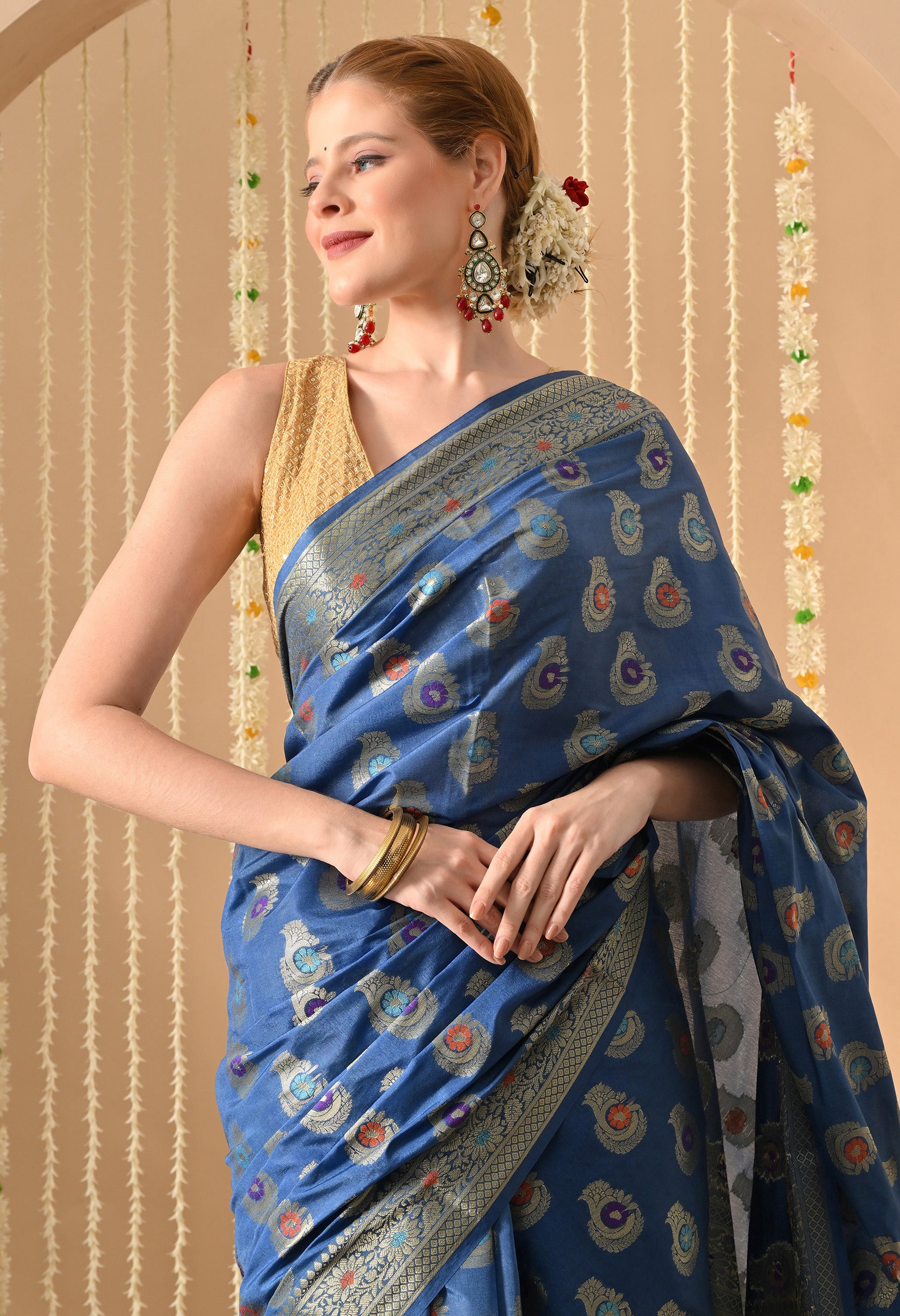 A close-up shot of a woman looking to the side, wearing a blue silk-blend Banarasi-style saree from The Weavers Shop by Raavi. The saree showcases colorful Tilfi work with metallic zari in a traditional buta pattern. She is styled with a sleeveless gold blouse, large earrings, and gold bangles, set against a background of hanging floral garlands.