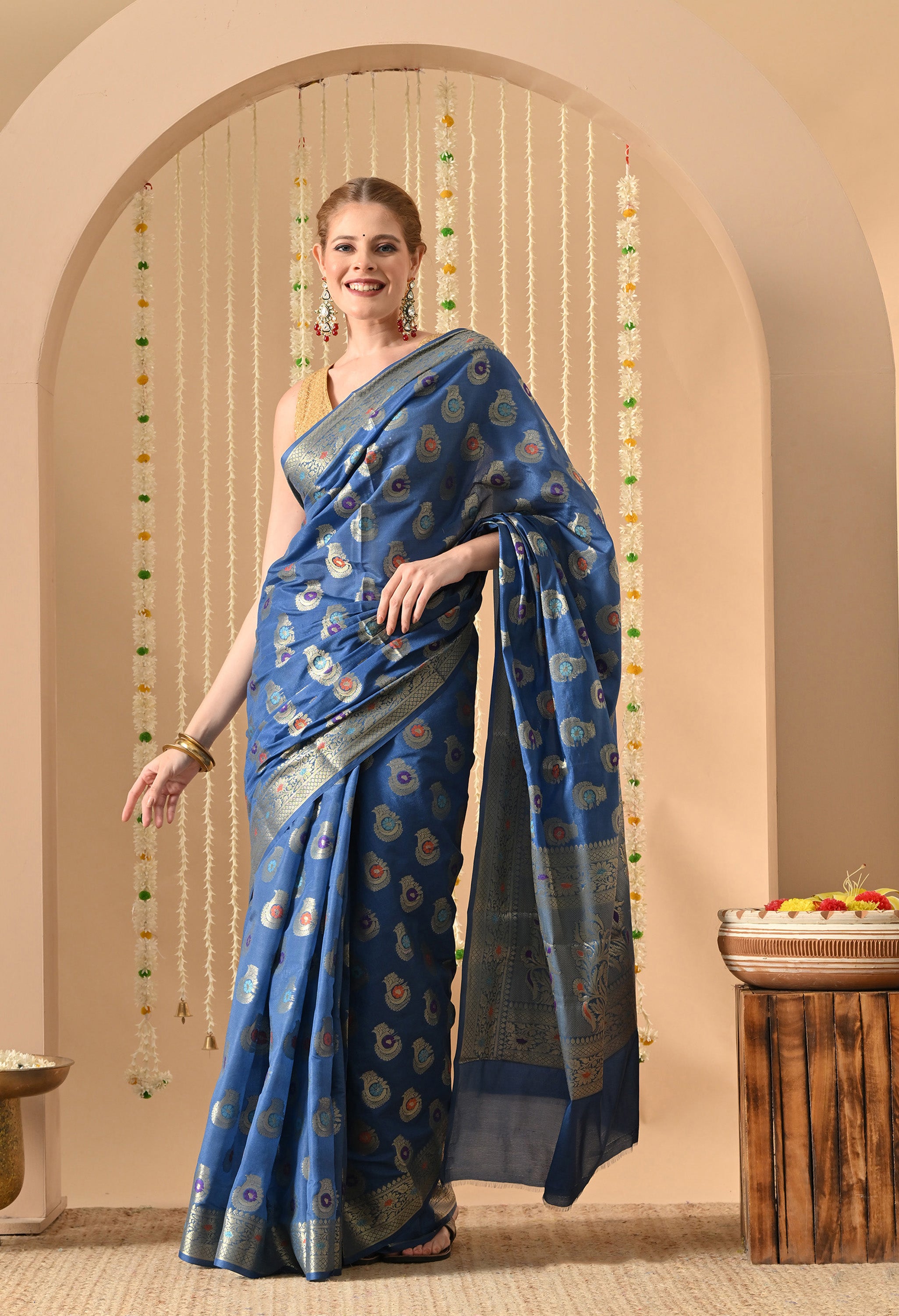 A woman models a vibrant blue silk saree from The Weavers Shop by Raavi, featuring intricate Tilfi and Zari work with multi-colored paisley and floral motifs. The saree is embellished with a dense metallic gold border and a decorative pallu. She is styled with a sleeveless gold sequined blouse, ornate red and green earrings, and gold bangles.