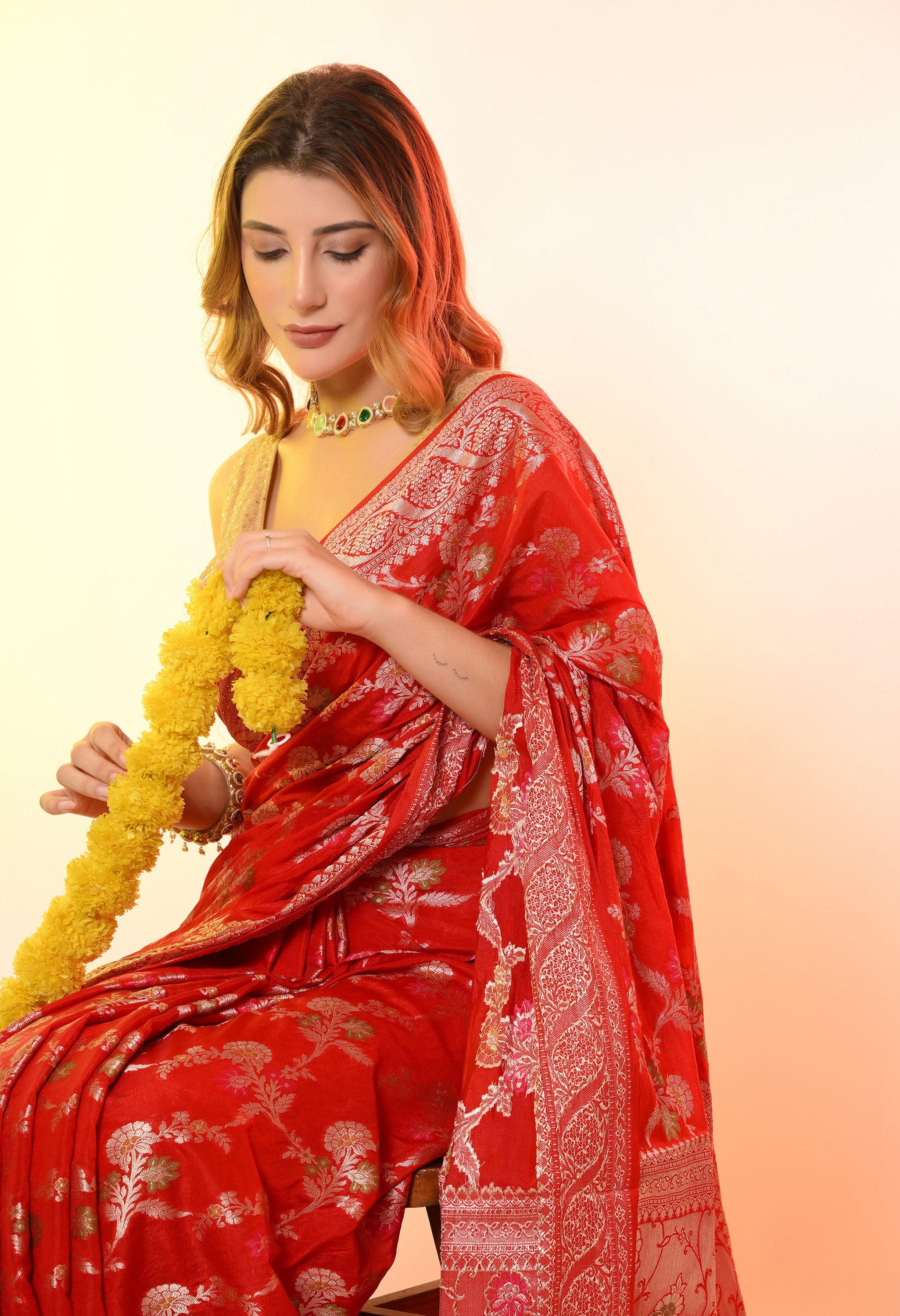 A woman sitting and holding a yellow flower garland while wearing a red Banarasi-style silk-blend saree by The Weavers Shop by Raavi. The shot provides a detailed view of the silver zari and floral Tilfi work on the machine-woven fabric. She is accessorized with a gold blouse, bangles, and a choker necklace.