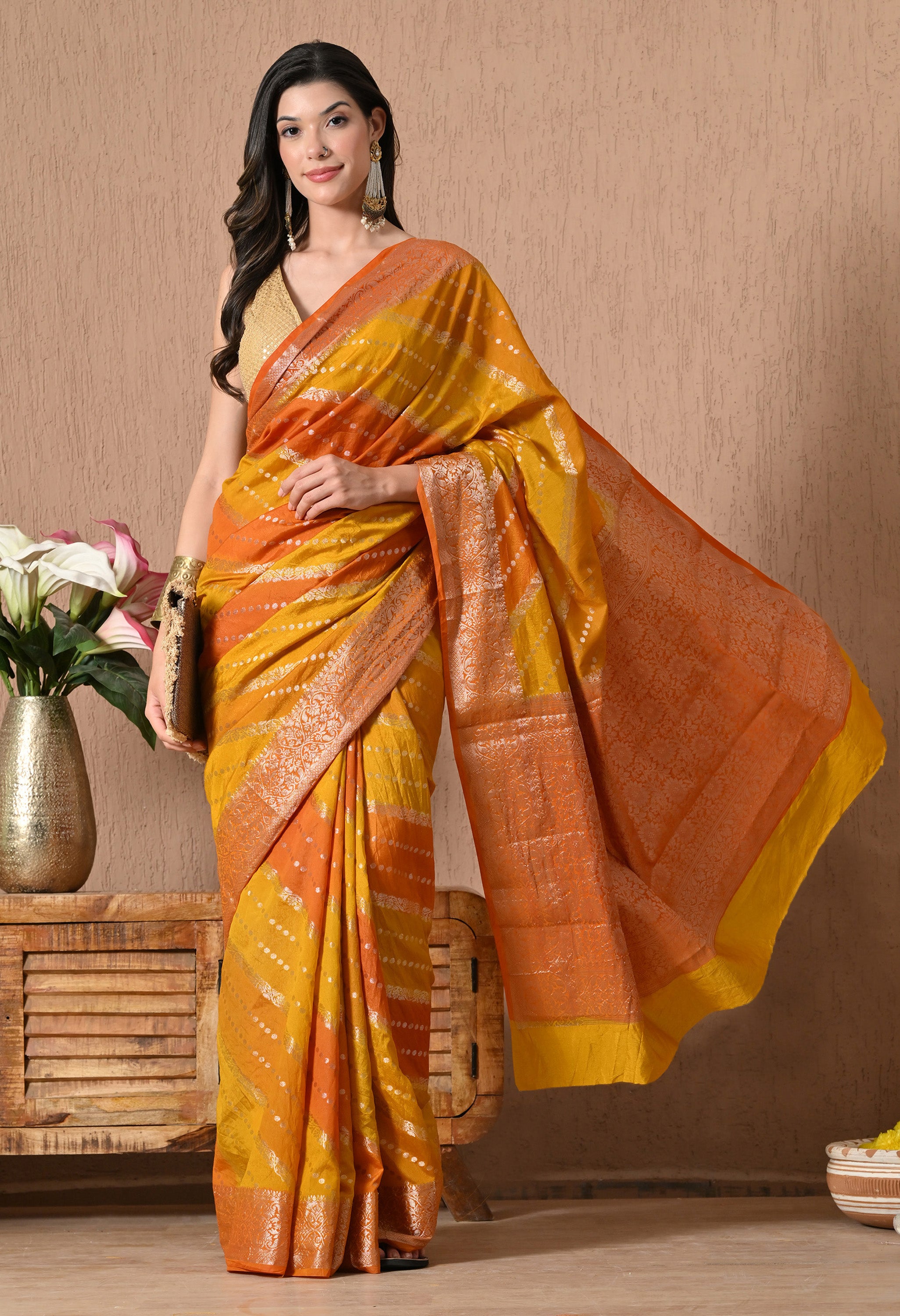 A woman poses in a vibrant Rangkat Banarasi silk saree from The Weavers Shop, featuring a sunset palette of mustard yellow and deep orange diagonal stripes. The saree is adorned with intricate golden zari work in floral patterns along the borders and pallu, complemented by delicate silver-toned polka dots throughout the body. She pairs the drape with a sleeveless gold blouse and traditional jewellery.