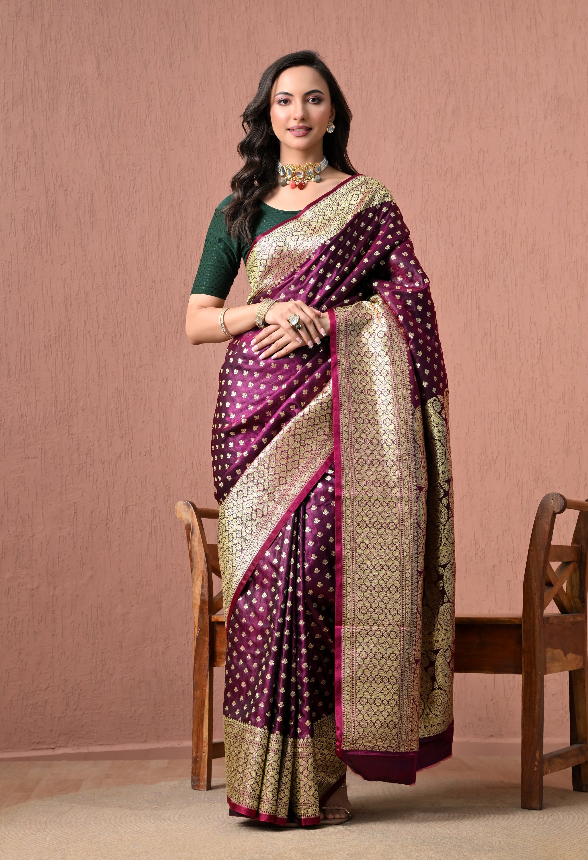 Full-length studio shot of a model wearing a luxurious deep wine-purple pure Katan silk Banarasi saree with intricate gold zari butis and a heavy gold border, paired with a green elbow-length blouse and a traditional choker necklace.
