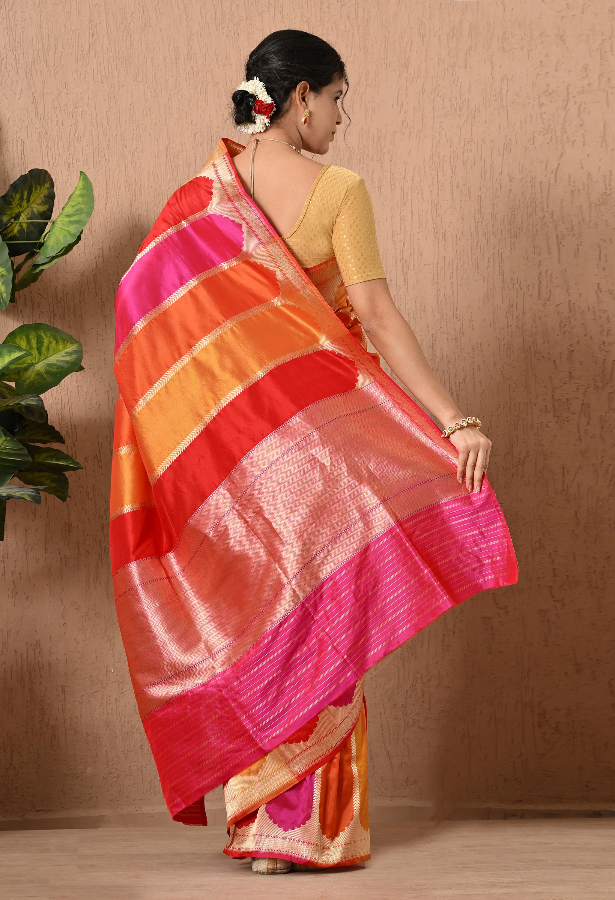 A rear view of a woman showcasing a handwoven Pure Katan Silk saree from The Weavers Shop by Raavi. The image highlights the traditional Banaras Rangkat weaving technique, featuring vibrant diagonal stripes in rich shades of pink, orange, and red. The saree is handwoven and is  adorned with intricate Zari work, including a wide, shimmering gold border with fine horizontal detailing. This Silk Mark Certified piece is paired with a gold  blouse and a floral hair arrangement.
