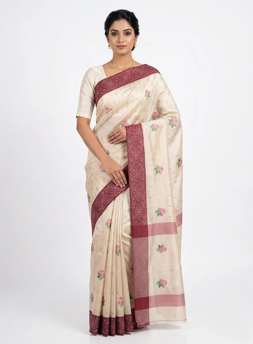 Elegant beige silk blend saree featuring pink and green floral embroidered butis. The saree is designed with a broad maroon border and pallu accented with intricate floral woven patterns and gold zari work.