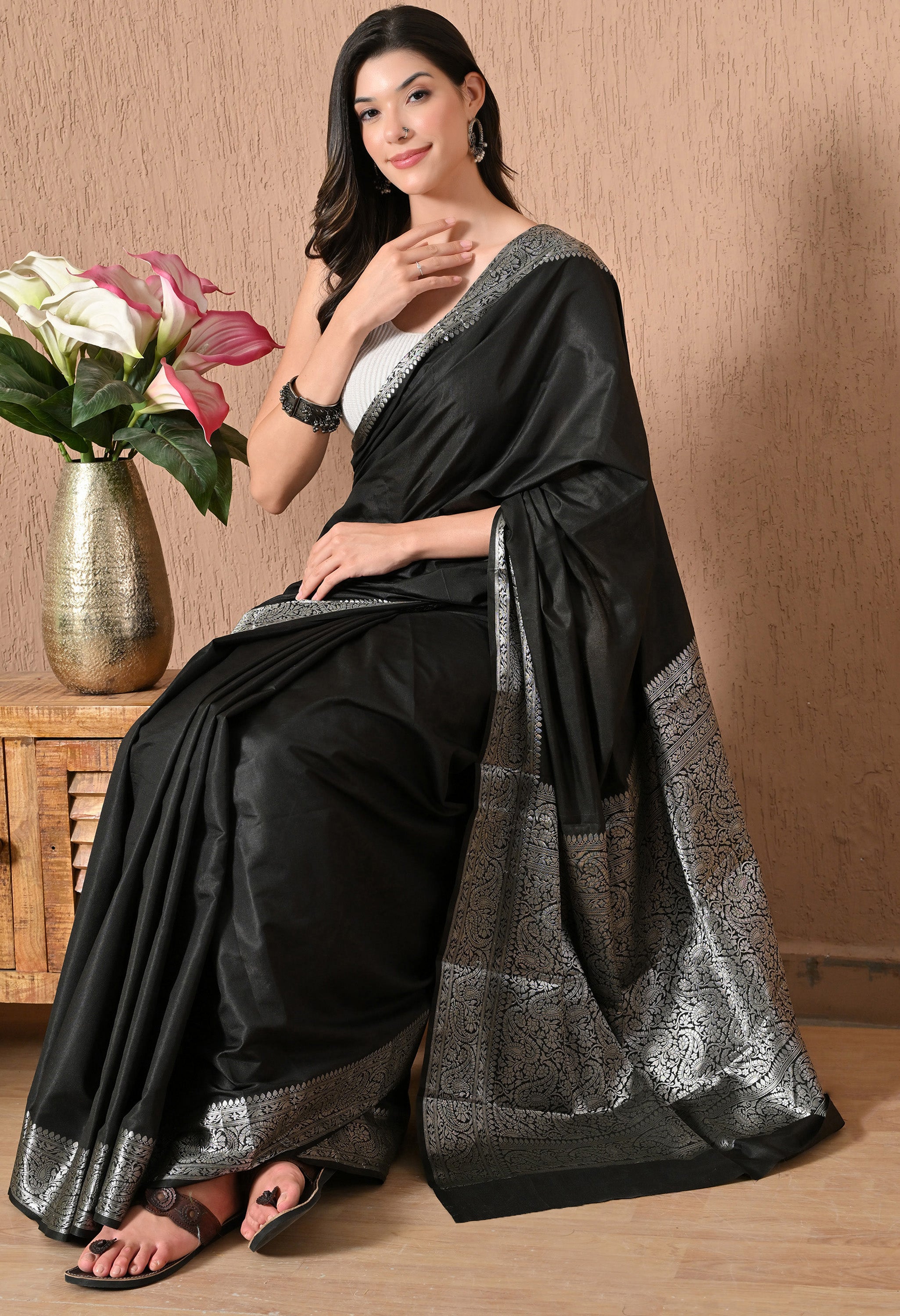 A seated model wearing a black silk saree from The Weavers Shop by Raavi, displaying the rich texture of the silk and how the silver zari border reflects light in an indoor setting.