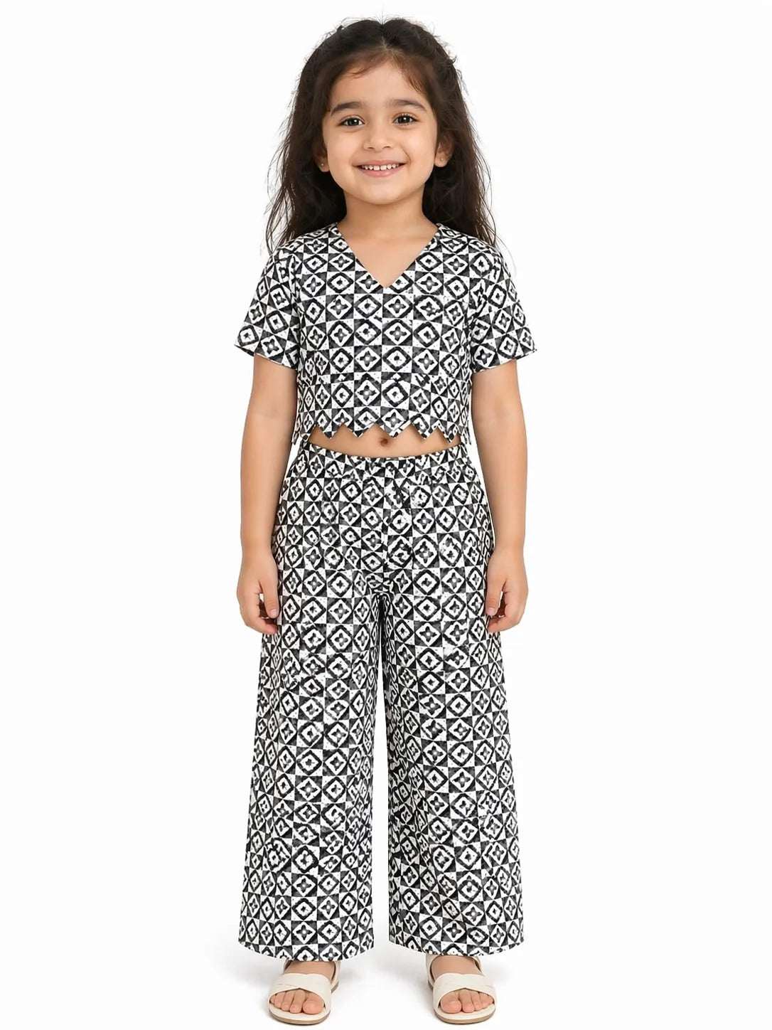 A young girl wearing a black and white geometric print ethnic co-ord set. The outfit features a V-neck, short-sleeved crop top with a decorative scalloped hemline and matching high-waisted wide-leg trousers, showcasing an all-over intricate patterned design.