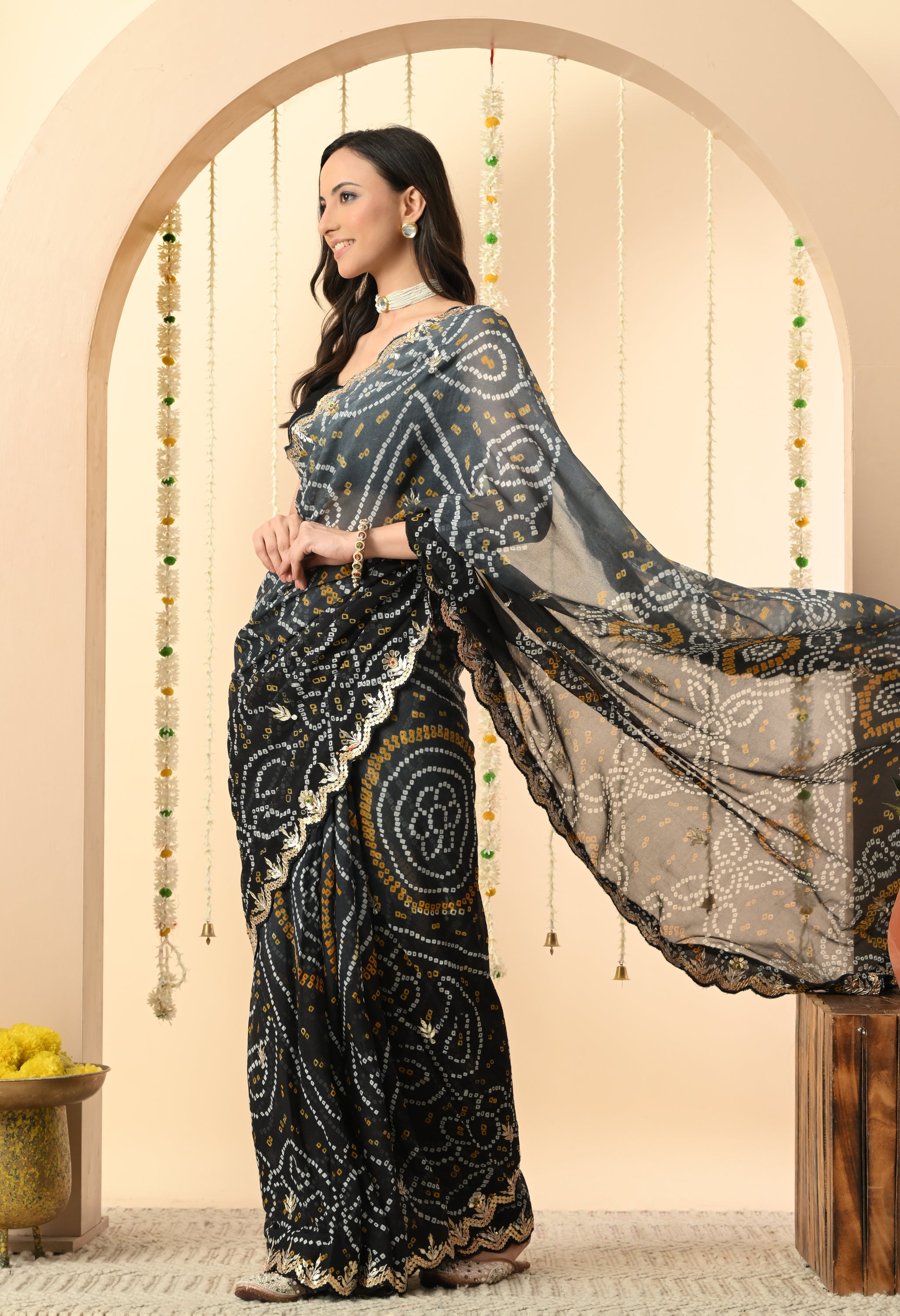 A woman stands in profile, looking toward the side with a smile to showcase a sophisticated black georgette saree from The Weavers Shop by Raavi. This handcrafted Rajasthani Bandhani saree, designed for a festive event or a classy evening party, features traditional white and yellow tie-dye patterns and an intricate gold Gota Patti scalloped border. She is styled with a sleeveless black blouse and a white pearl choker, posed against a neutral-toned archway with the saree's pallu elegantly draped and flowing