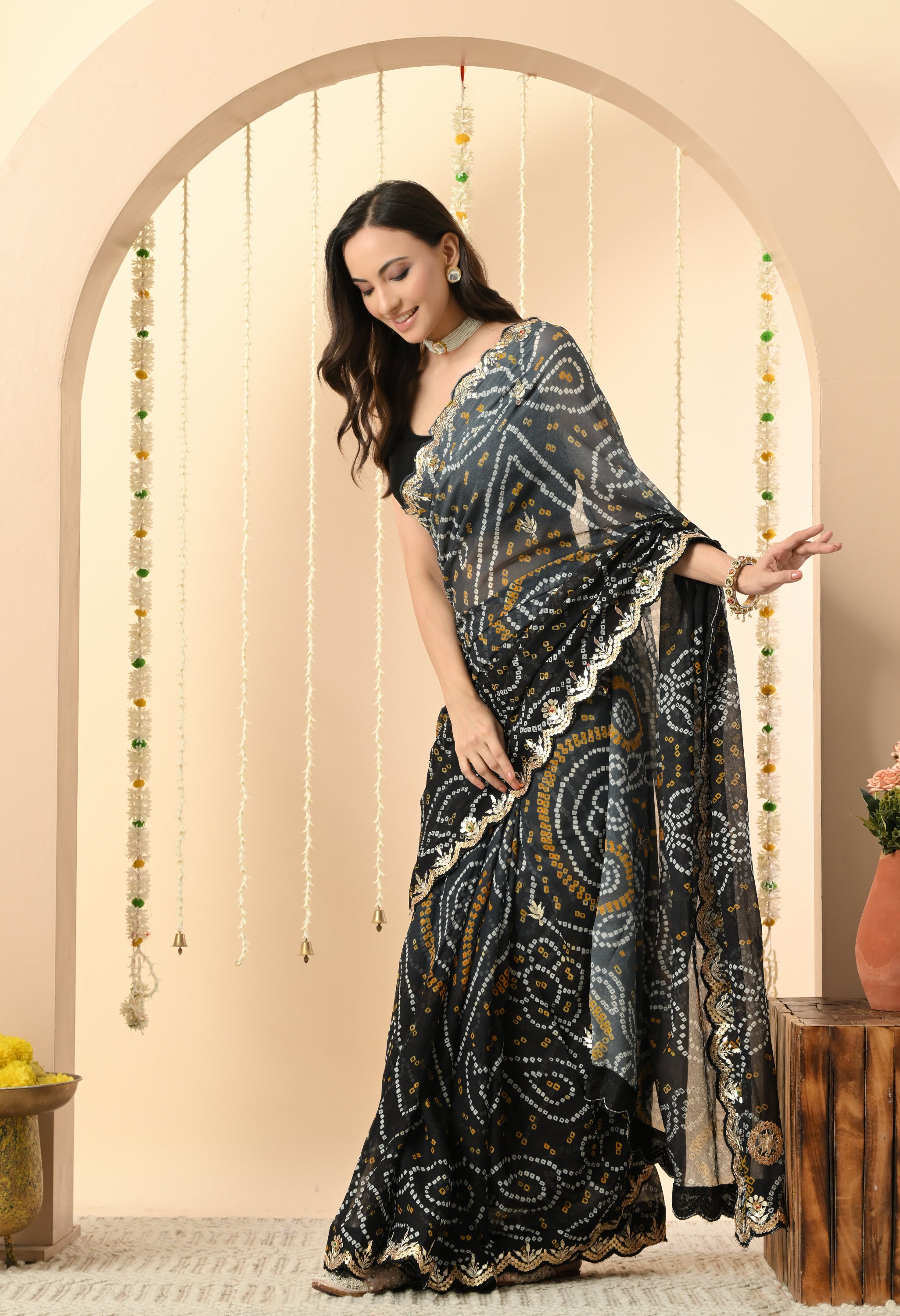 A woman is looking down with a gentle smile, showcasing a luxurious black georgette saree from The Weavers Shop by Raavi. This handcrafted Rajasthani Bandhani saree, ideal for a festive event or a high-end party, features intricate white and mustard yellow tie-dye patterns and a striking gold Gota Patti scalloped border. She is styled with a sleeveless black blouse and a white pearl choker, posed in a festive setting decorated with hanging jasmine garlands.