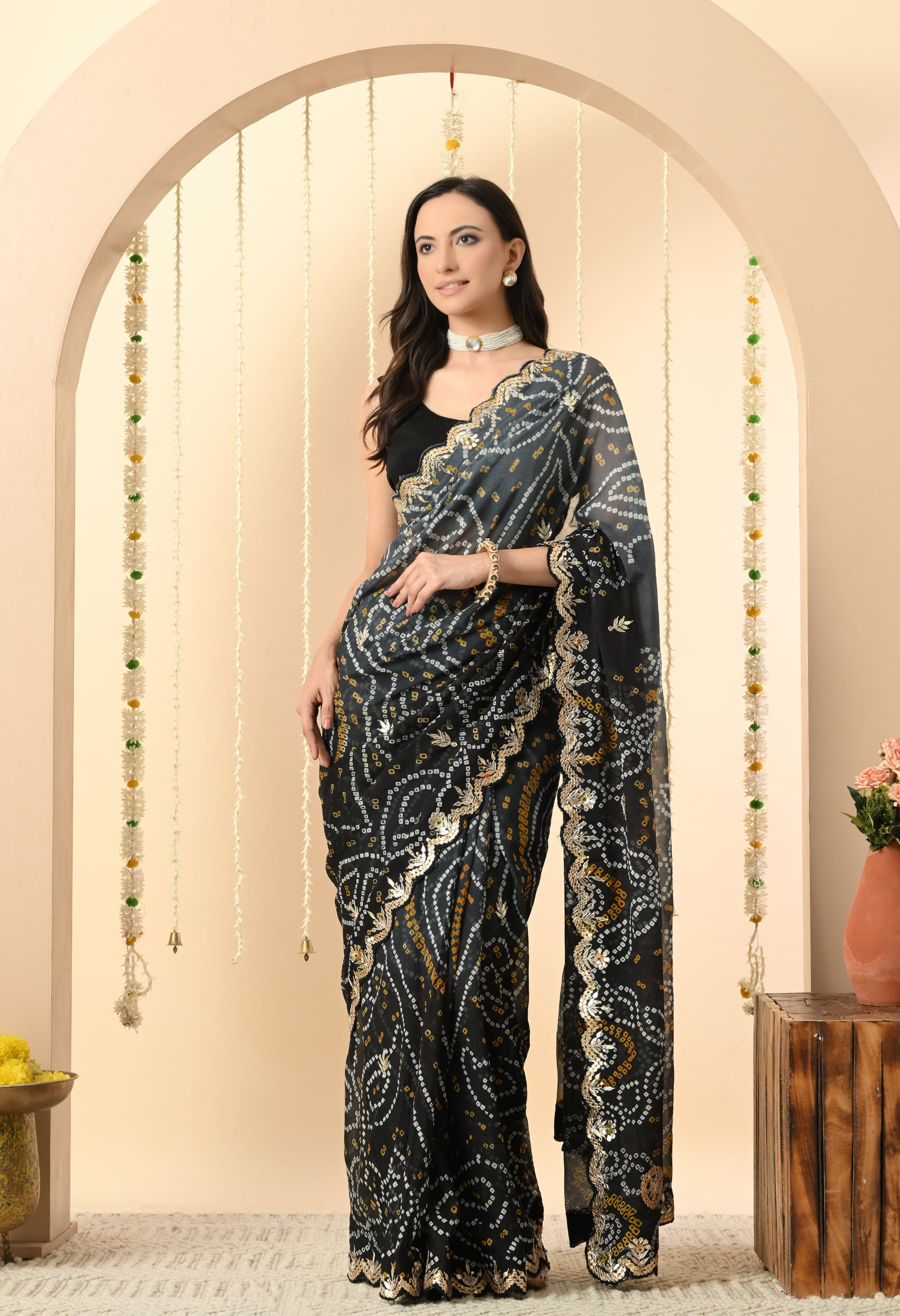 A woman stands elegantly wearing a black georgette saree from The Weavers Shop by Raavi, designed for a festive event or wedding. This handcrafted Rajasthani Bandhani saree features traditional white and yellow tie-dye patterns and is adorned with intricate gold Gota Patti work along the scalloped borders. She is styled with a sleeveless black blouse and a white pearl choker, posed against a neutral-toned archway decorated with jasmine garlands.