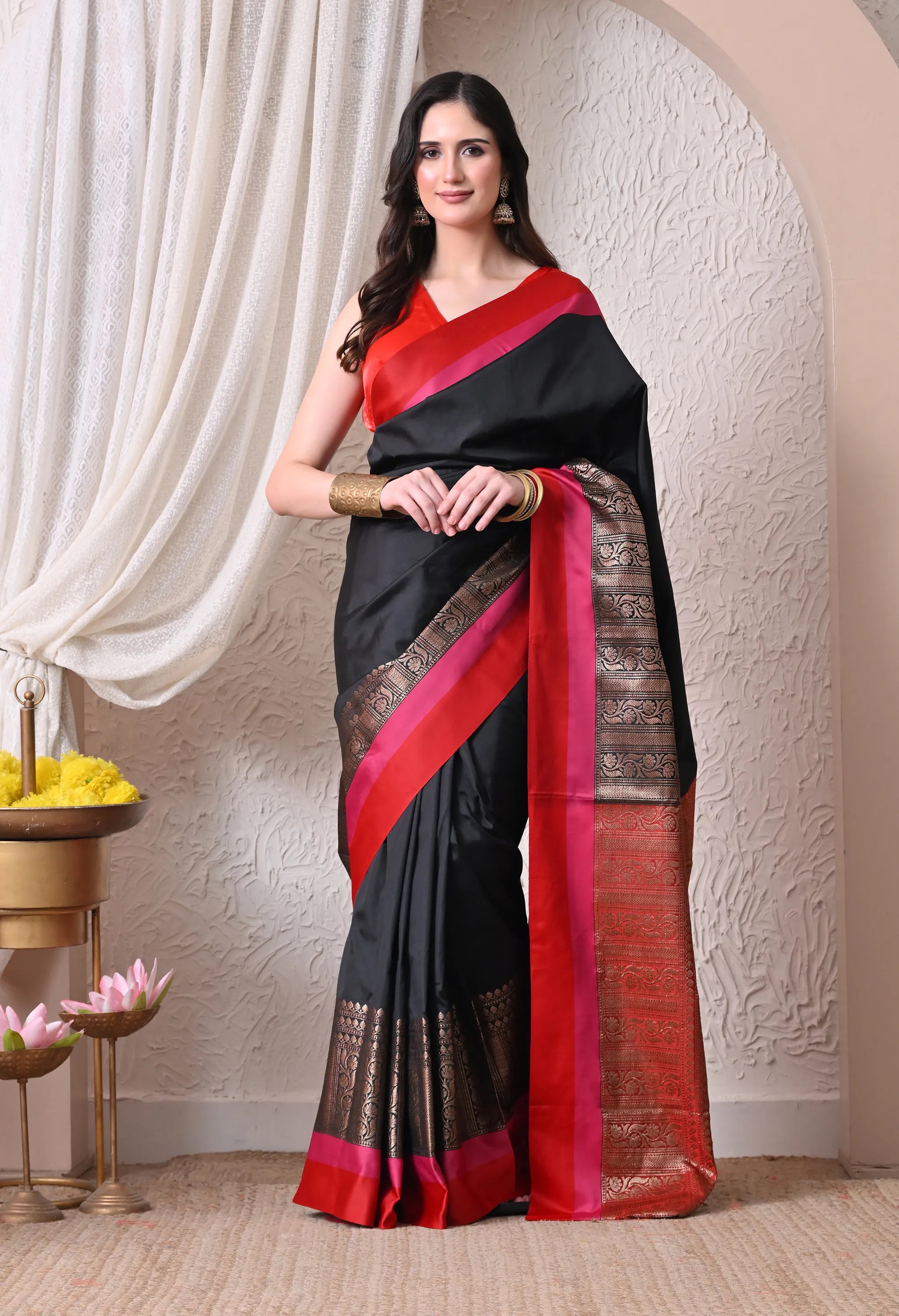 A woman posing in a sleek black silk blend saree featuring a distinct pink and red border with antique gold zari work
