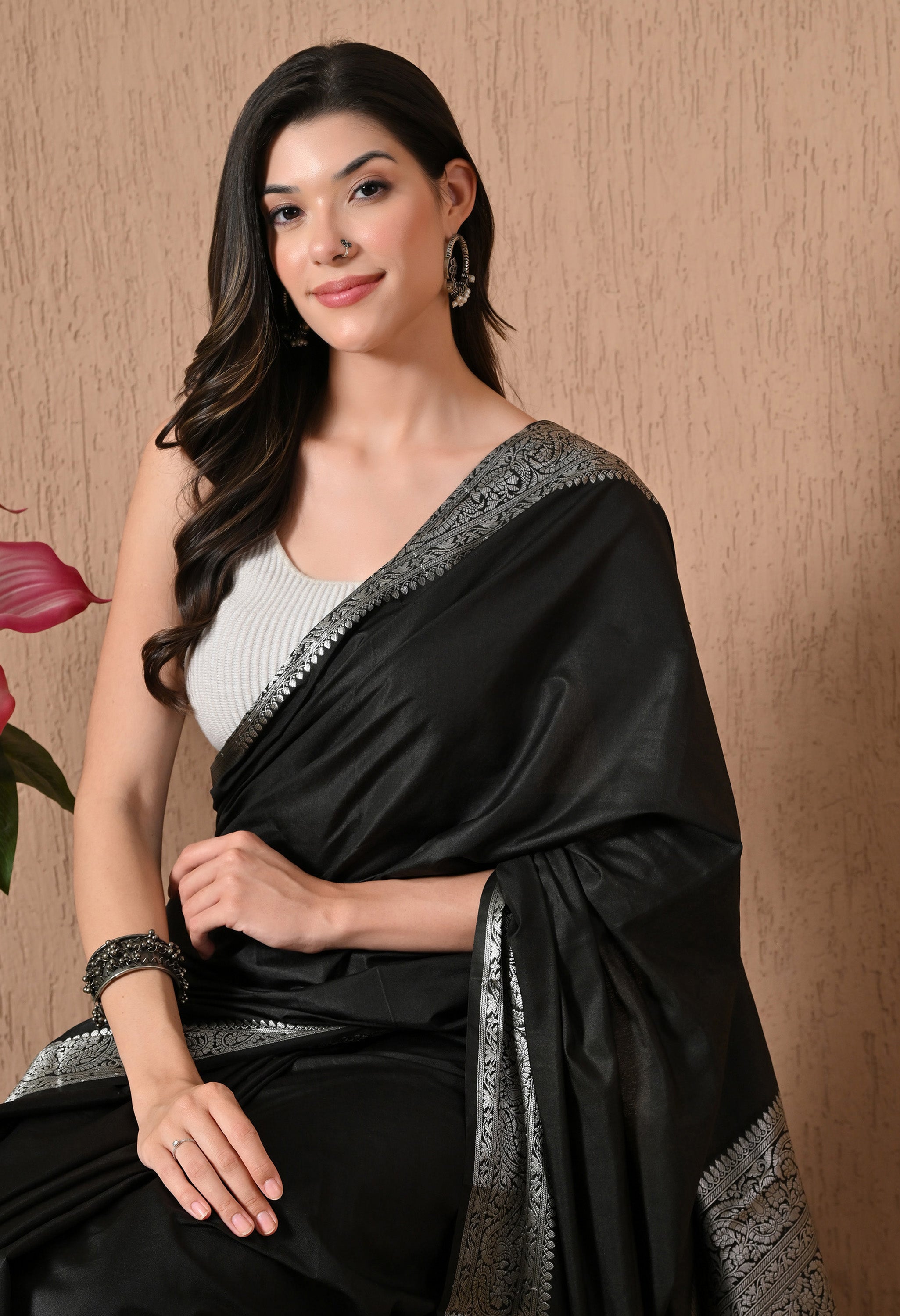 A portrait-style image of a woman in a black silk saree by The Weavers Shop by Raavi, highlighting the elegant pairing of a white sleeveless blouse with traditional silver jhumka earrings and a nose stud.