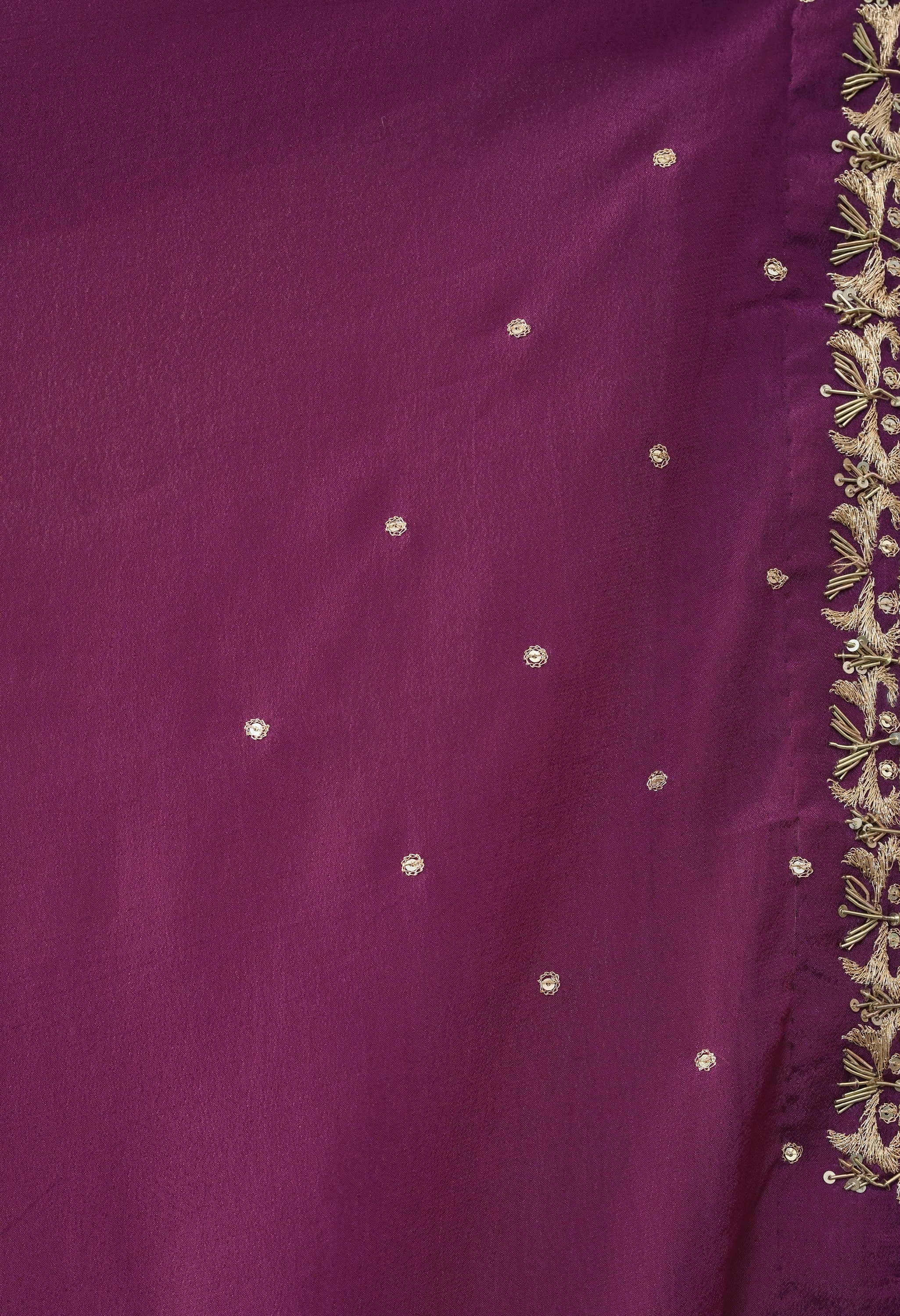 A detailed close-up of the blouse piece of the wine-coloured crepe saree from The Weavers Shop by Raavi, highlighting the intricate gold zardozi hand-embroidery along the border. The embroidery features fine metallic threadwork and small sequins in a repeating floral motif. Scattered across the rich purple crepe fabric are delicate, individual gold sequence dots, showcasing the craftsmanship of the hand-worked design.