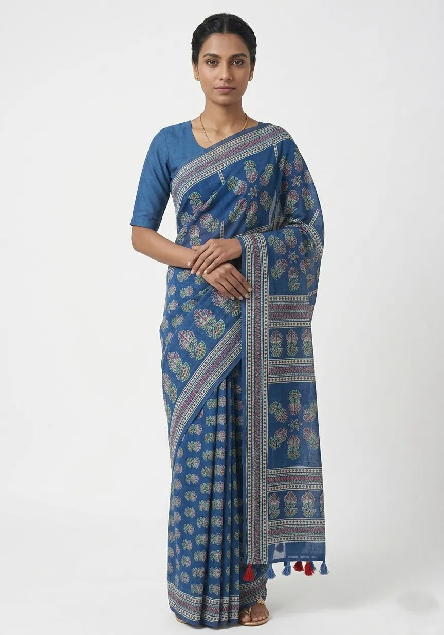 A woman modeling a deep blue indigo cotton saree featuring traditional floral prints, a decorative patterned border, and colorful tassels on the pallu.
