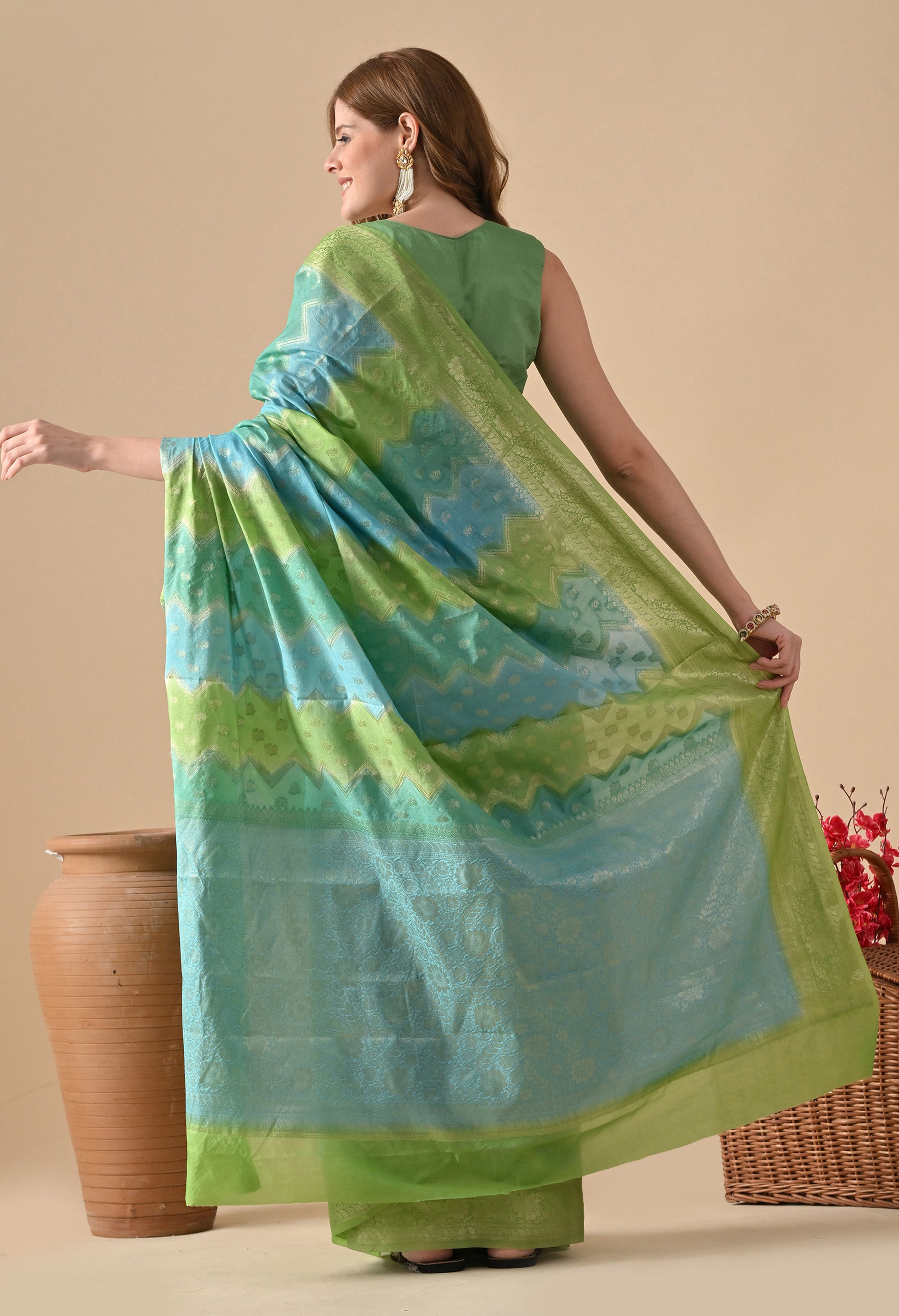 A back view of a woman showcasing a Banarasi Rangkat saree from The Weavers Shop. The image highlights the horizontal zigzag "Rangkat" color-blocking technique in alternating bands of lime green and turquoise blue. Detailed gold zari work is visible throughout the body and across the wide pallu. The woman is wearing a matching sleeveless green blouse and pearl-drop earrings, posing with her back to the camera to display the drape of the saree against a neutral studio background with a large clay vase and a 