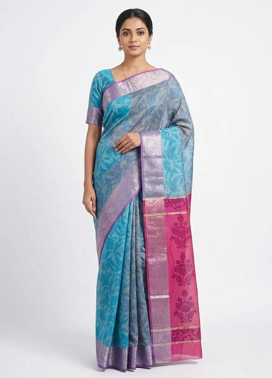 A woman posing in a blue-grey Banarasi silk saree with intricate silver woven motifs and a contrasting pink and gold-toned zari border.