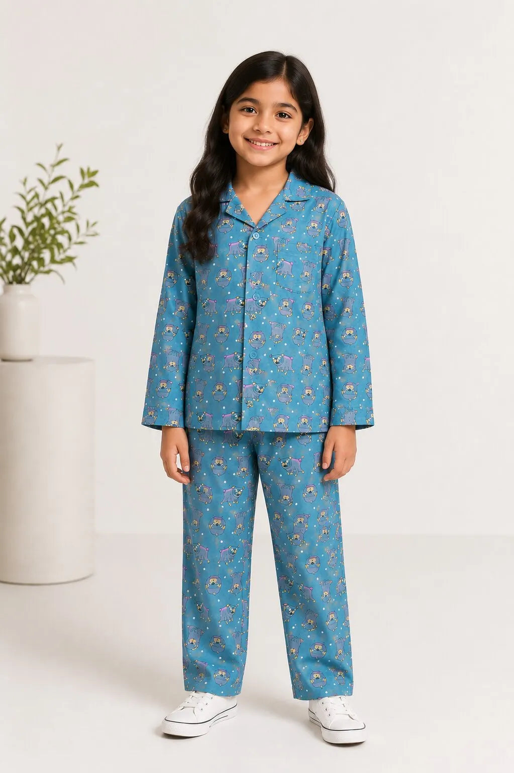 A young girl smiling in a bedroom, wearing a teal blue cotton night suit with a grey hedgehog print and holding a teddy bear.