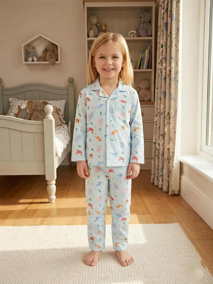 A young girl standing in a sunlit bedroom wearing a light blue satin night suit with a playful all-over animal print. She is smiling in a cozy setting featuring a classic white bed and a soft cream-colored rug.