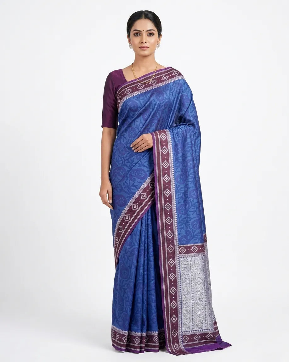 An Indian woman posing in a royal blue silk blend saree featuring tonal floral patterns, a purple geometric border, and a silver-toned pallu. She is wearing a solid purple blouse and a thin gold chain against a white background.
